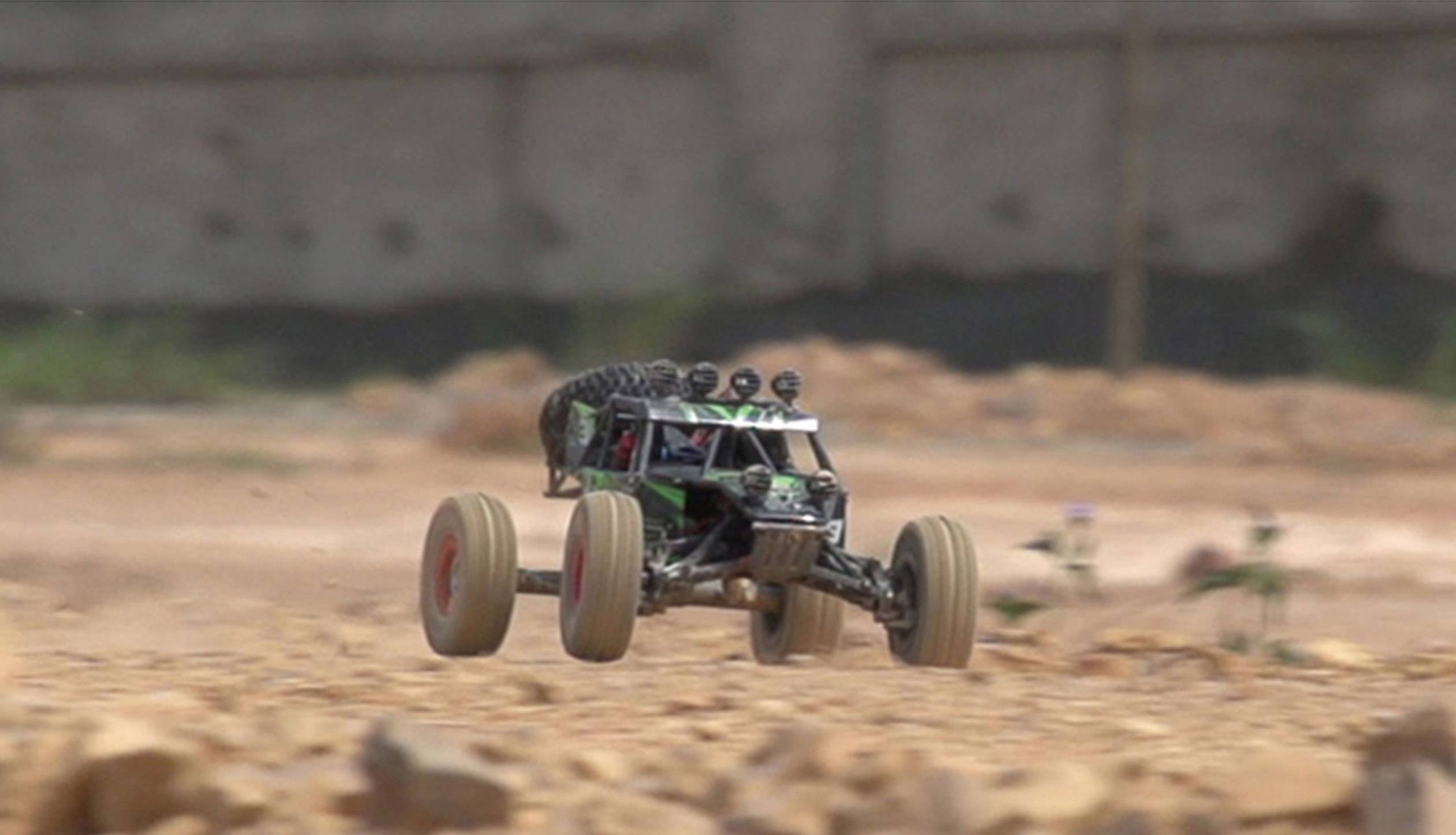 Ein ferngesteuertes Auto fährt mit hoher Geschwindigkeit über unebenes, steiniges Gelände. Im Hintergrund ist eine unscharfe Mauer erkennbar.