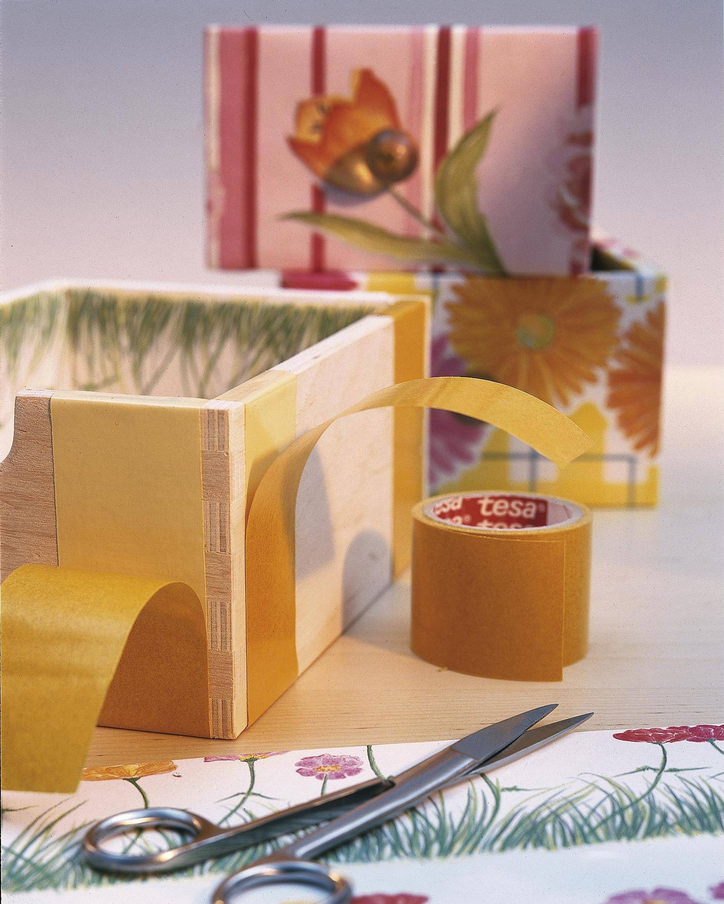 Geschenkboxen mit Blumenmustern, gelbes Klebeband und eine Schere auf einem Tisch. Das Arrangement zeigt Bastelbedarf für Geschenkverpackungen.