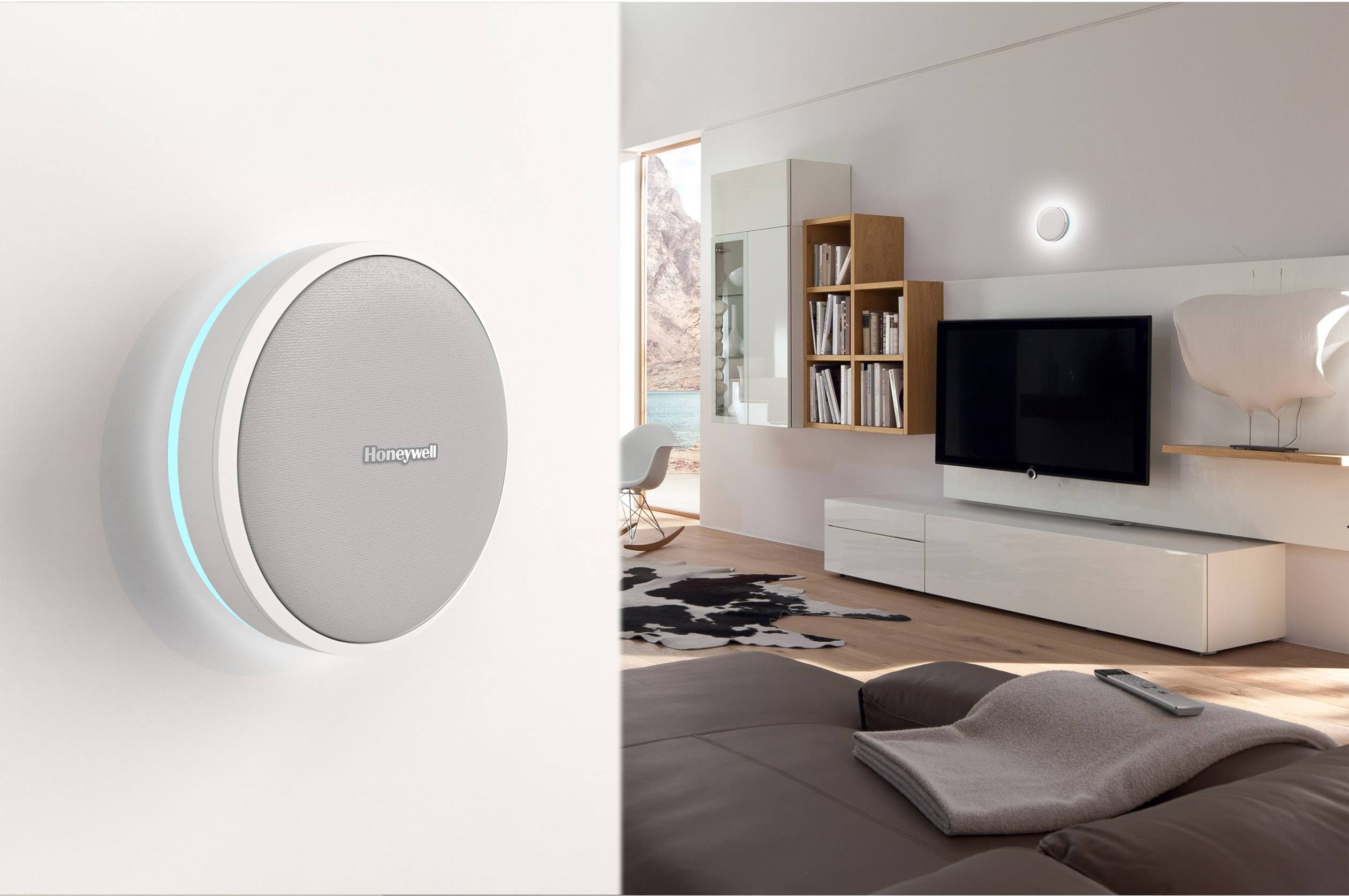 Ein modernes Wohnzimmer mit grauem Sofa, Fernseher und Regalen. An der Wand links ein rundes, silbernes Smart-Home-Gerät.