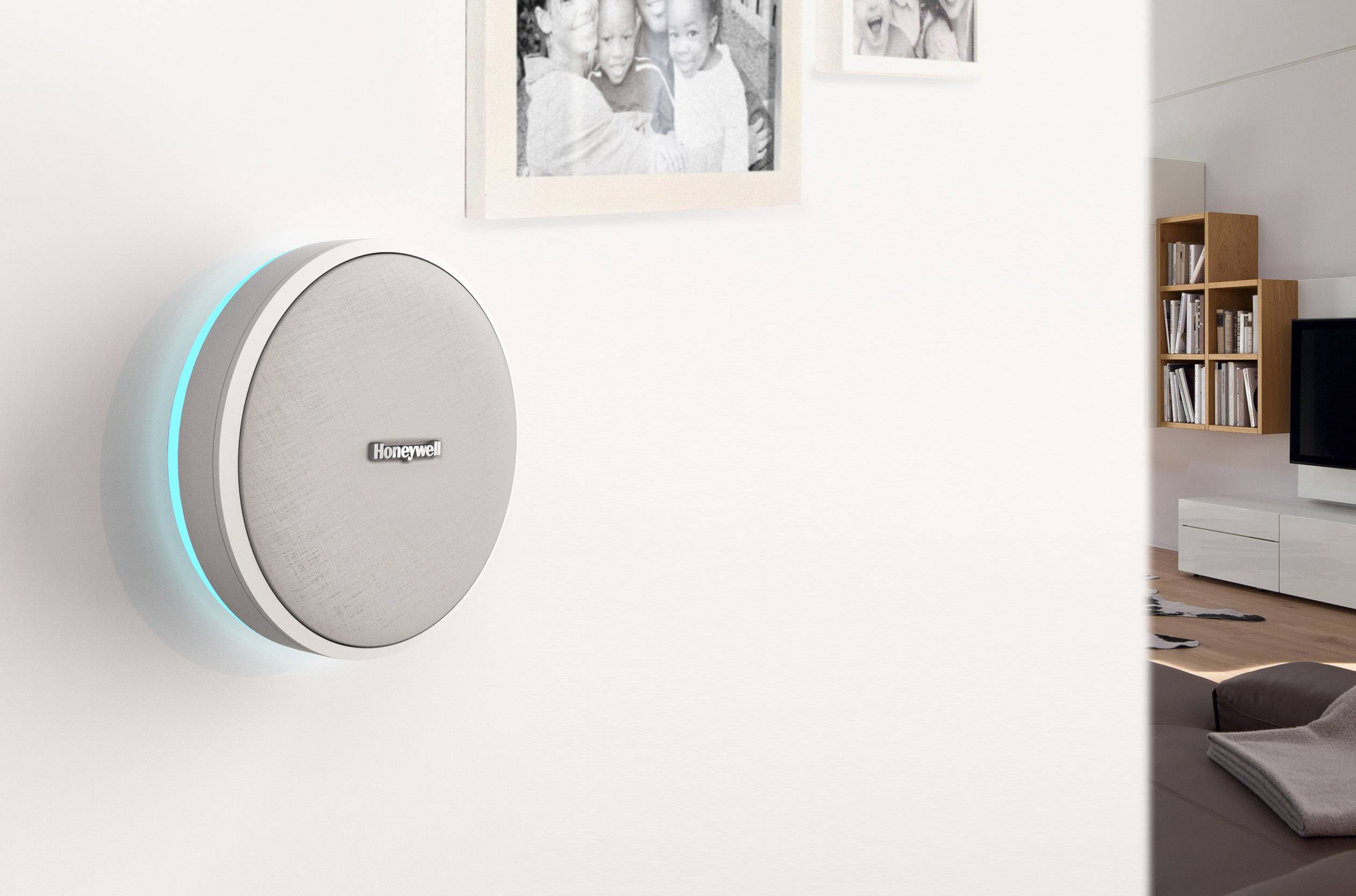 Smart-Thermostat an der Wand eines Wohnzimmers, daneben zwei Bilderrahmen. Wohnzimmermöbel und Fernseher im Hintergrund.