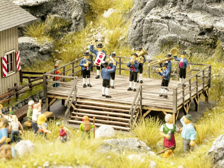 Eine kleine Blaskapelle spielt auf einer erhöhten hölzernen Plattform in einer Miniaturlandschaft, umgeben von Figuren und Felsen.