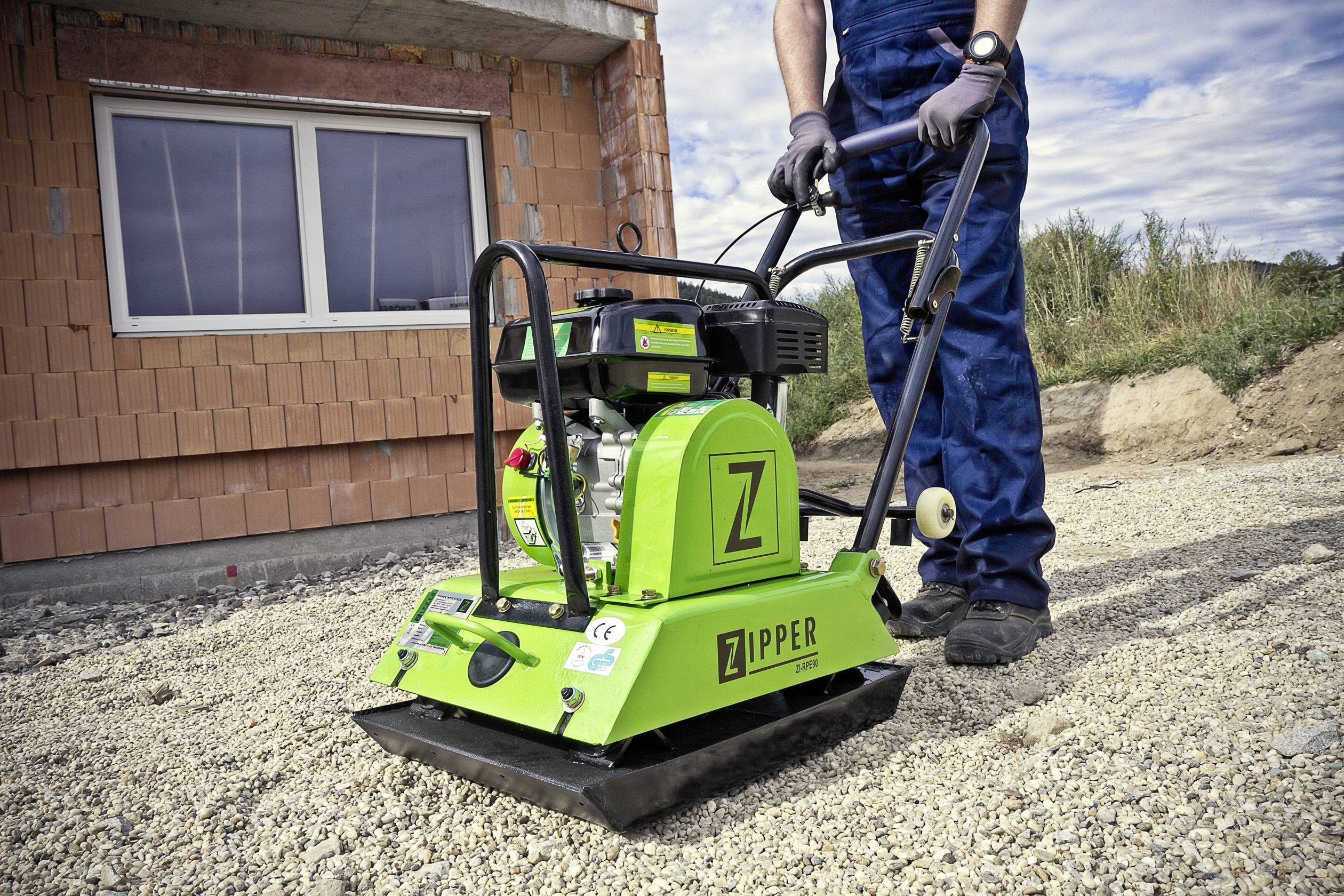 Person in Arbeitskleidung bedient eine grüne Rüttelplatte auf einer Schotterfläche vor einem Rohbau.