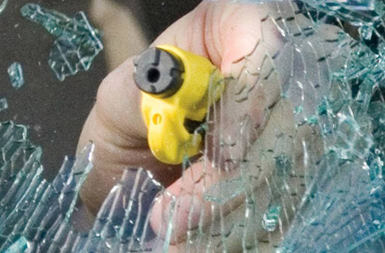 Hand hält gelbes Werkzeug, das durch zerbrochenes Glas stößt, um ein Fenster zu durchbrechen. Sicherheits- oder Notfallszene.