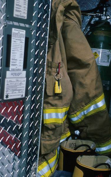 Feuerwehruniform hängt an der Wand neben Ausrüstungsgegenständen. Daneben ist ein Behälter mit Gefahrensymbolen zu sehen.