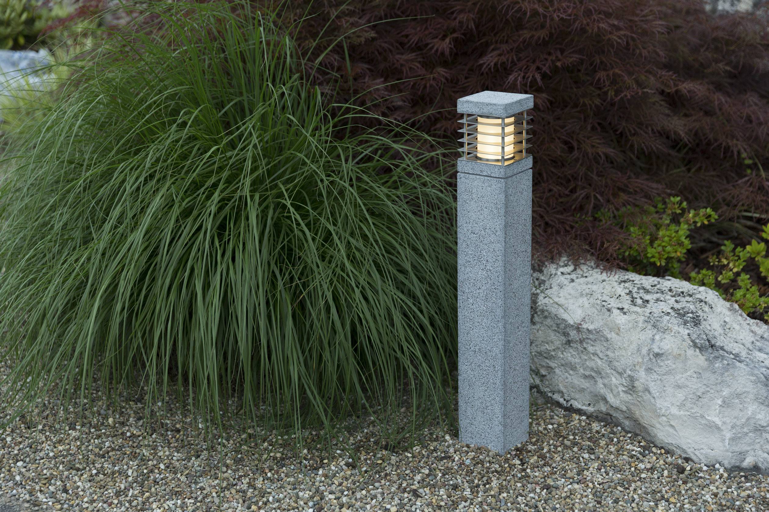 Eine Gartensäule mit eingeschaltetem Licht steht neben dekorativer Bepflanzung und großen Steinen auf einem Kiesbett.