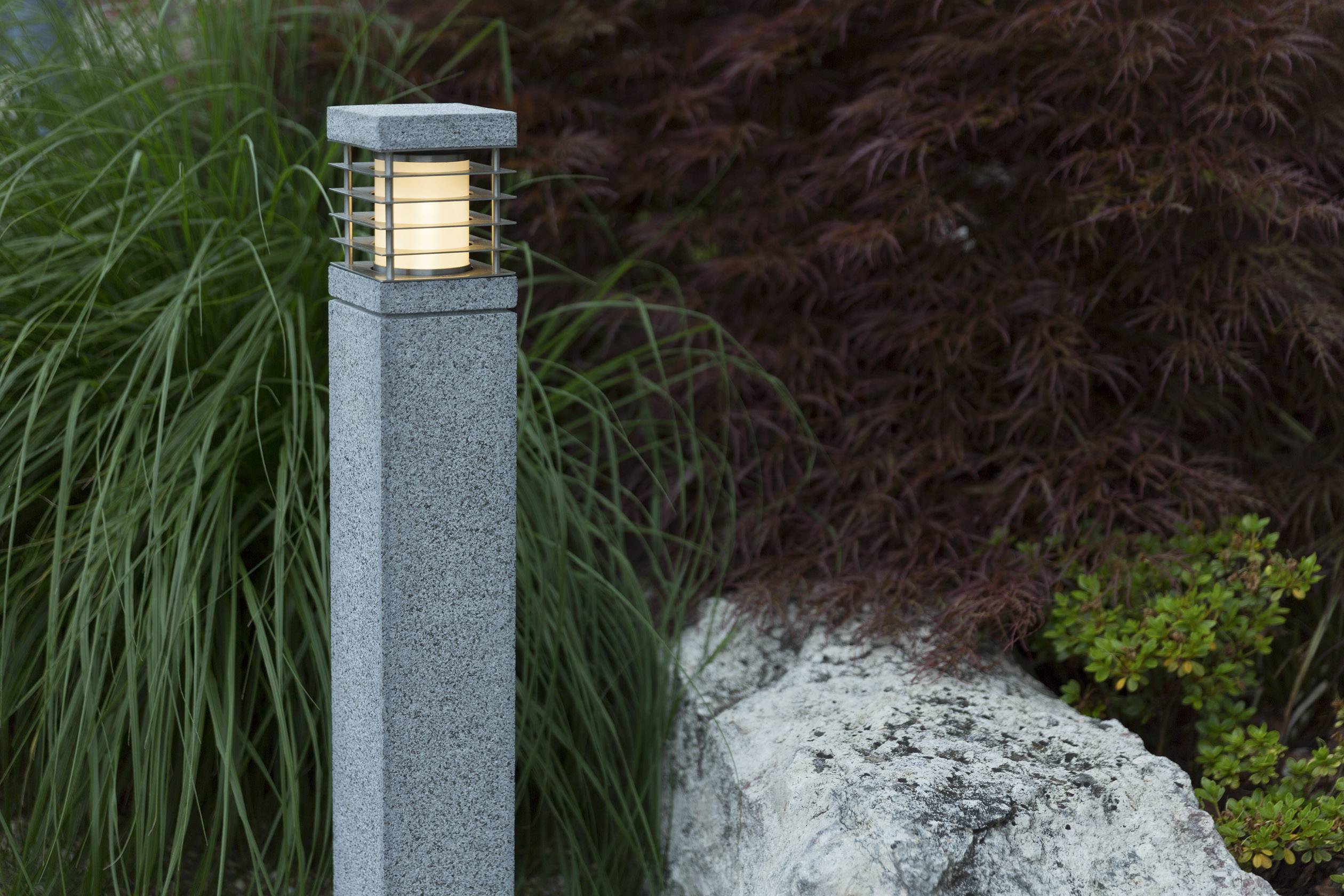 Eine moderne Stehlampe aus Stein leuchtet in einem Garten, umgeben von dekorativen Pflanzen und einem großen Felsen.