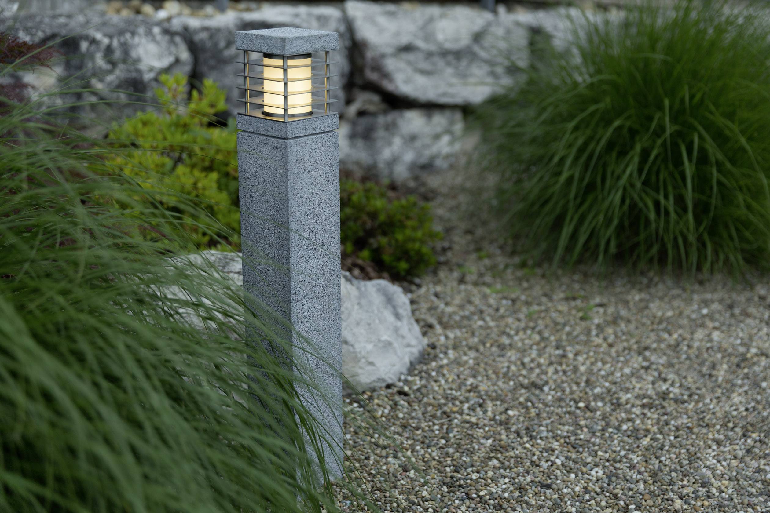 Eine moderne Steinlaterne leuchtet in einem Garten mit Kiesboden. Umgeben von grünen Gräsern und Steinen, schafft sie eine ruhige Atmosphäre.