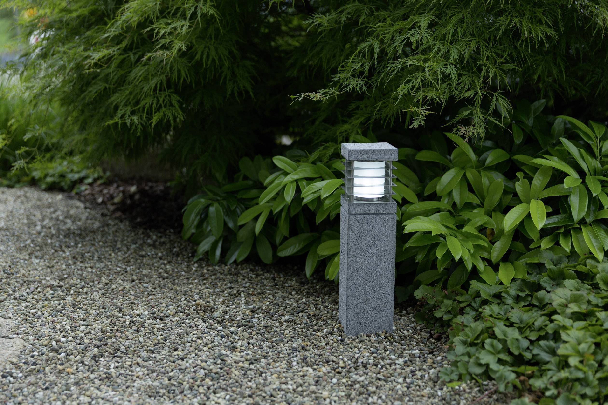 Außenleuchte auf einem Kiesweg, umgeben von grünen Pflanzen, spendet Licht in einem Gartenbereich.