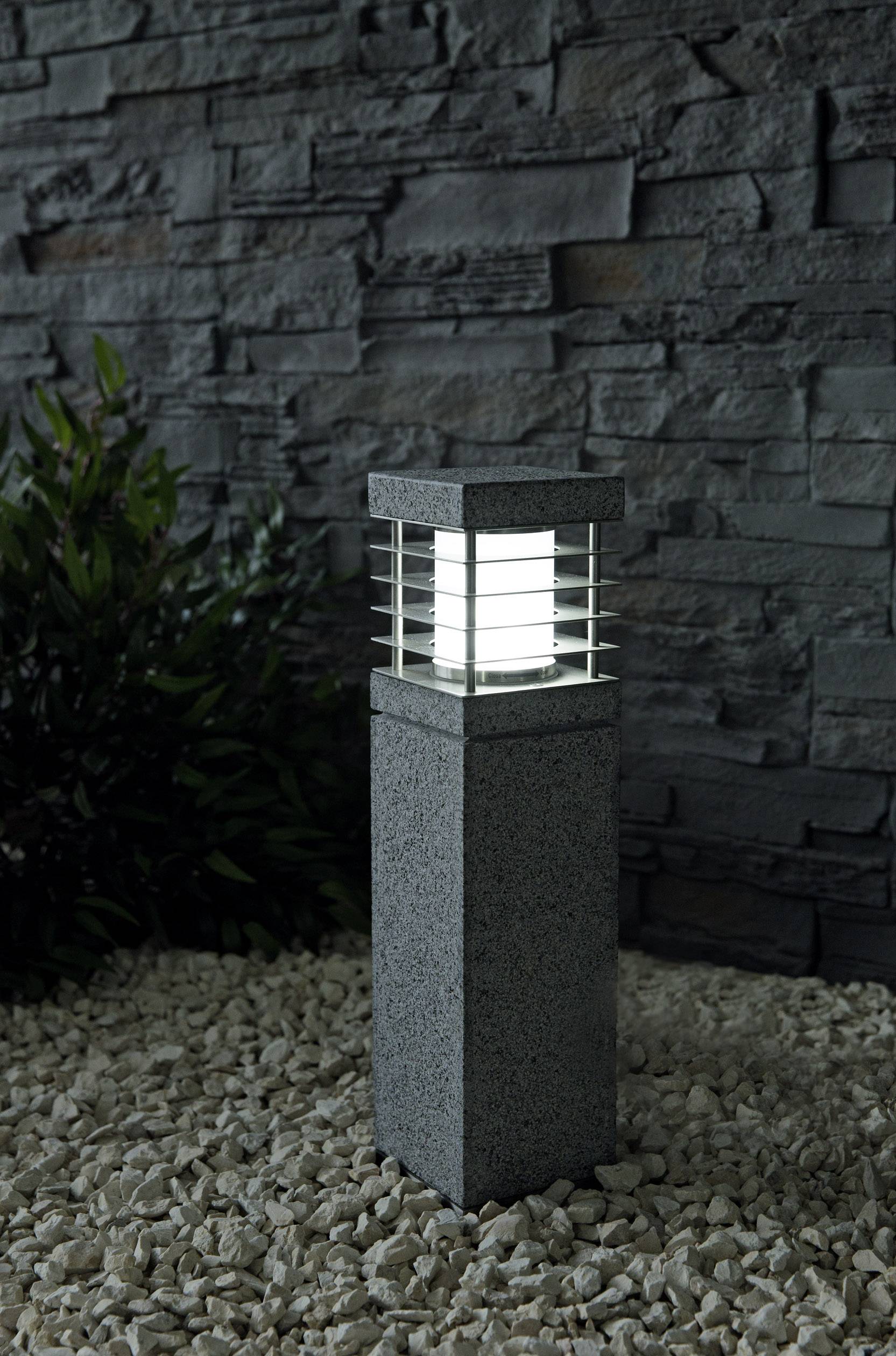 Steinlaterne auf Kies vor einer Steinmauer, von Pflanzen umgeben, bei Nacht beleuchtet.