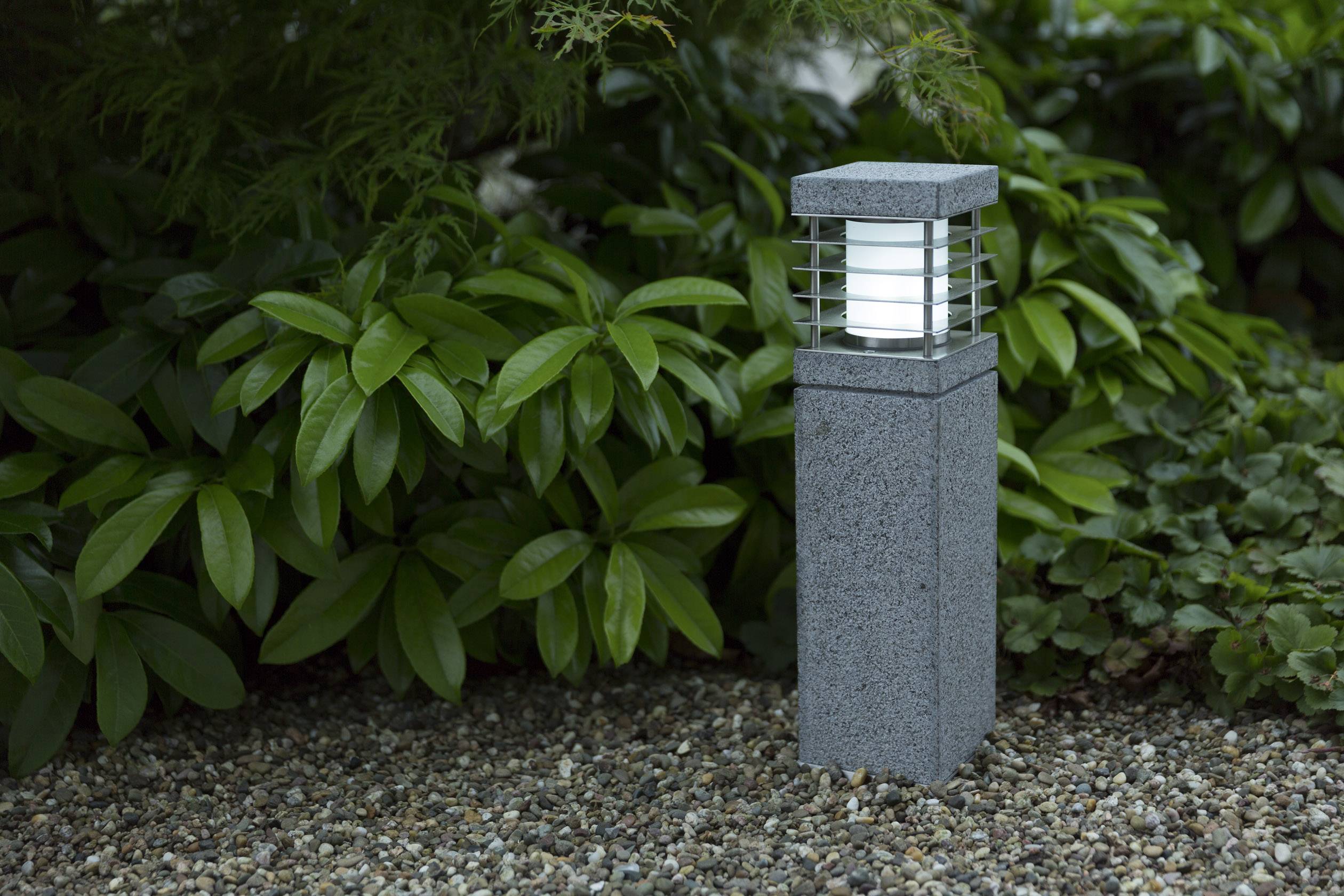 Eine steinerne Gartenlaterne steht auf einem Kiesweg, umgeben von grünen Blättern, und leuchtet sanft in der Dämmerung.