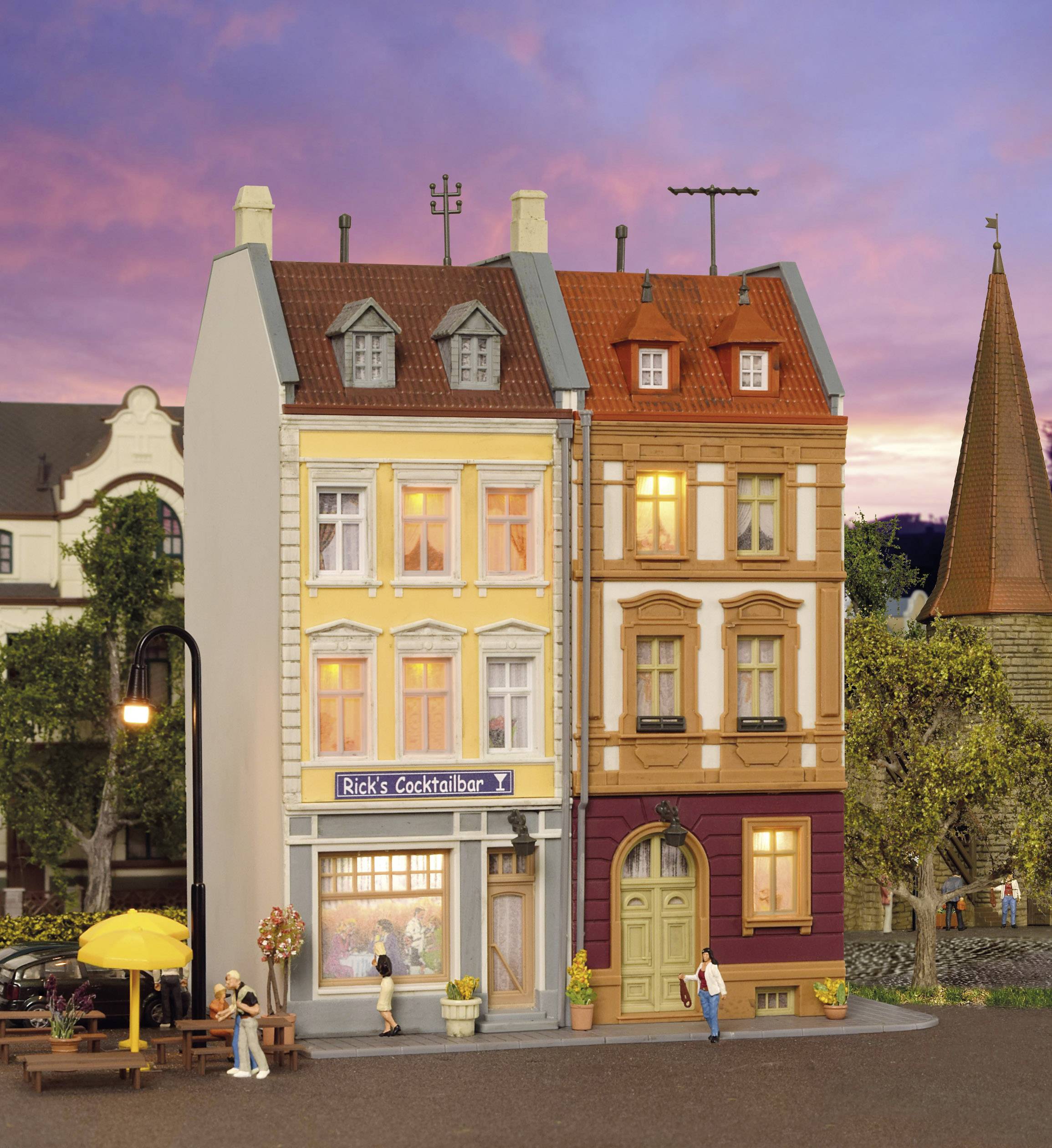 Zwei benachbarte, bunt gestrichene Gebäude mit einem kleinen Café im Erdgeschoss. Menschen sitzen draußen, während die Abenddämmerung einsetzt.