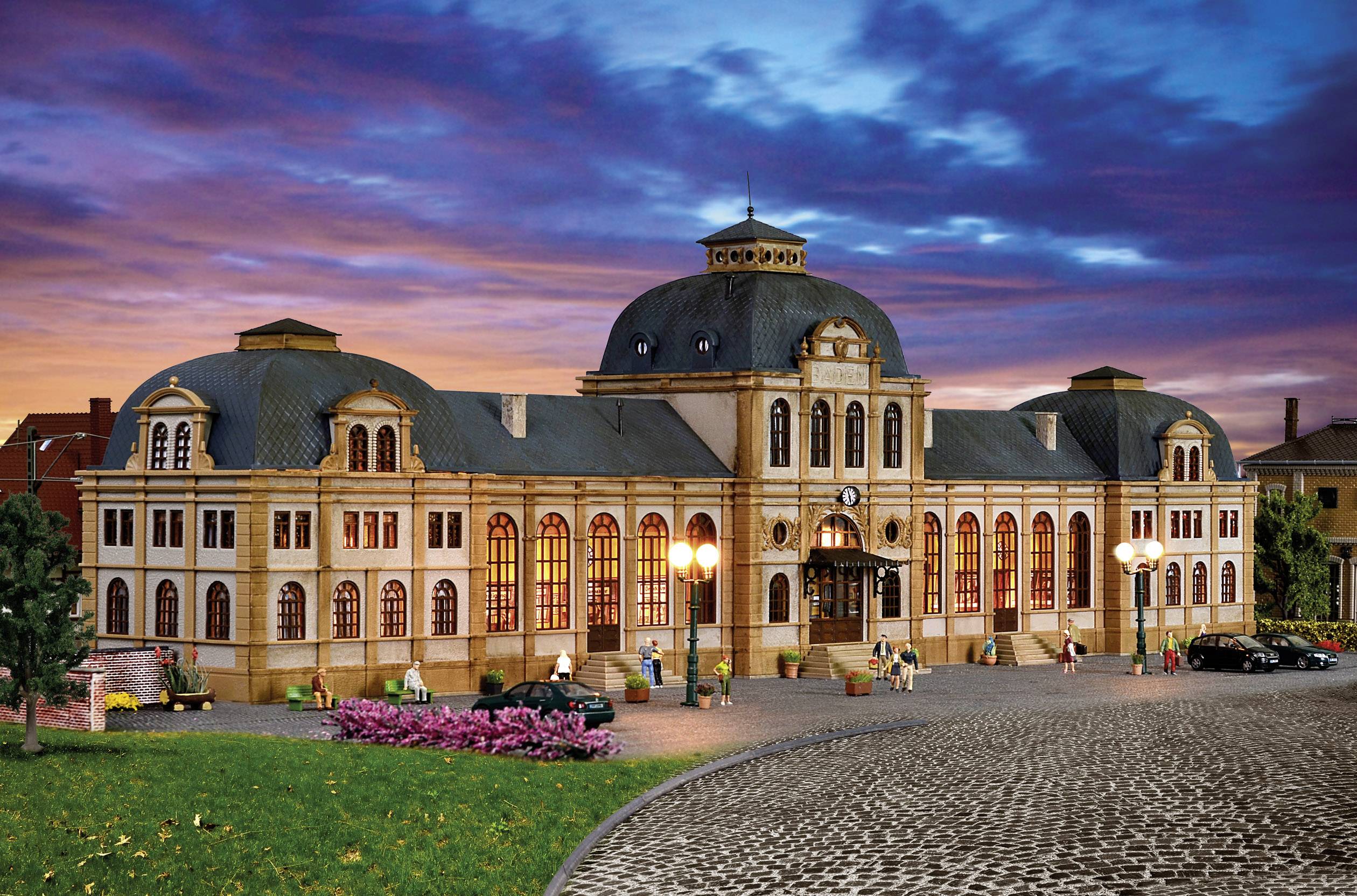 Ein großes, historisches Gebäude mit Kuppeldach und beleuchteten Fenstern bei Sonnenuntergang. Menschen und Autos vor dem Gebäude.