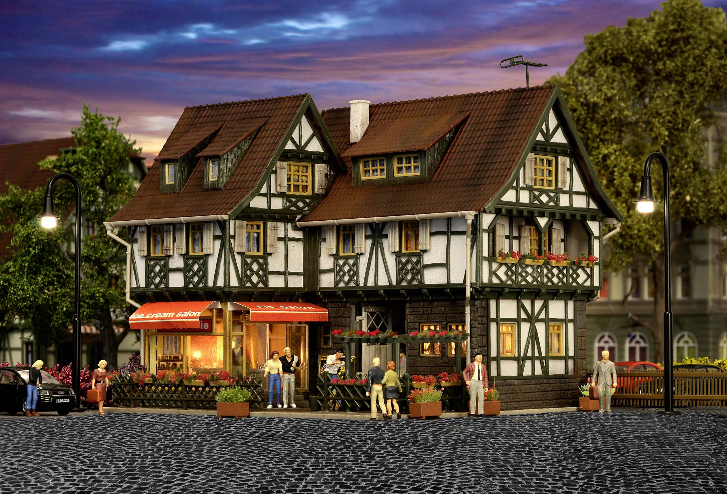 Fachwerkhaus mit Blumen, Laternen und Menschen auf Kopfsteinpflasterstraße bei Sonnenuntergang; ein Café im Erdgeschoss dient Gästen.