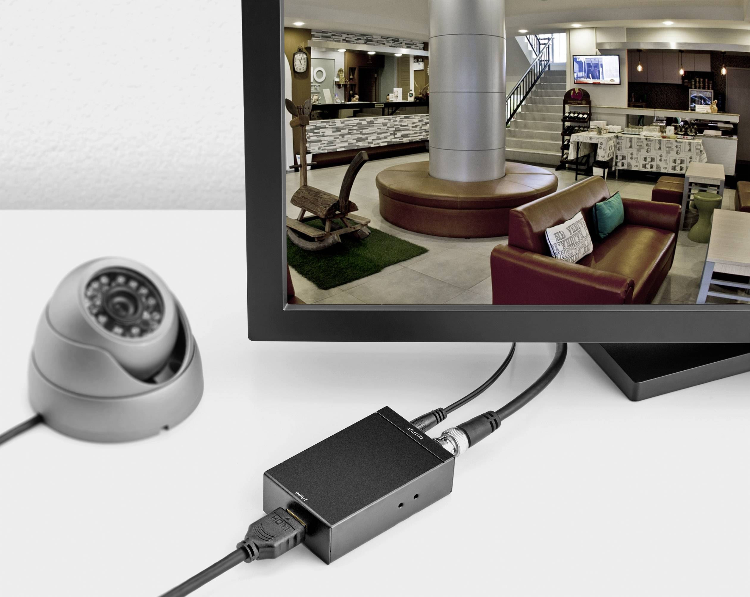 Überwachungsbildschirm zeigt Lobby eines Gebäudes. Kamera und Empfänger im Vordergrund. Moderne, gemütliche Einrichtung erkennbar.