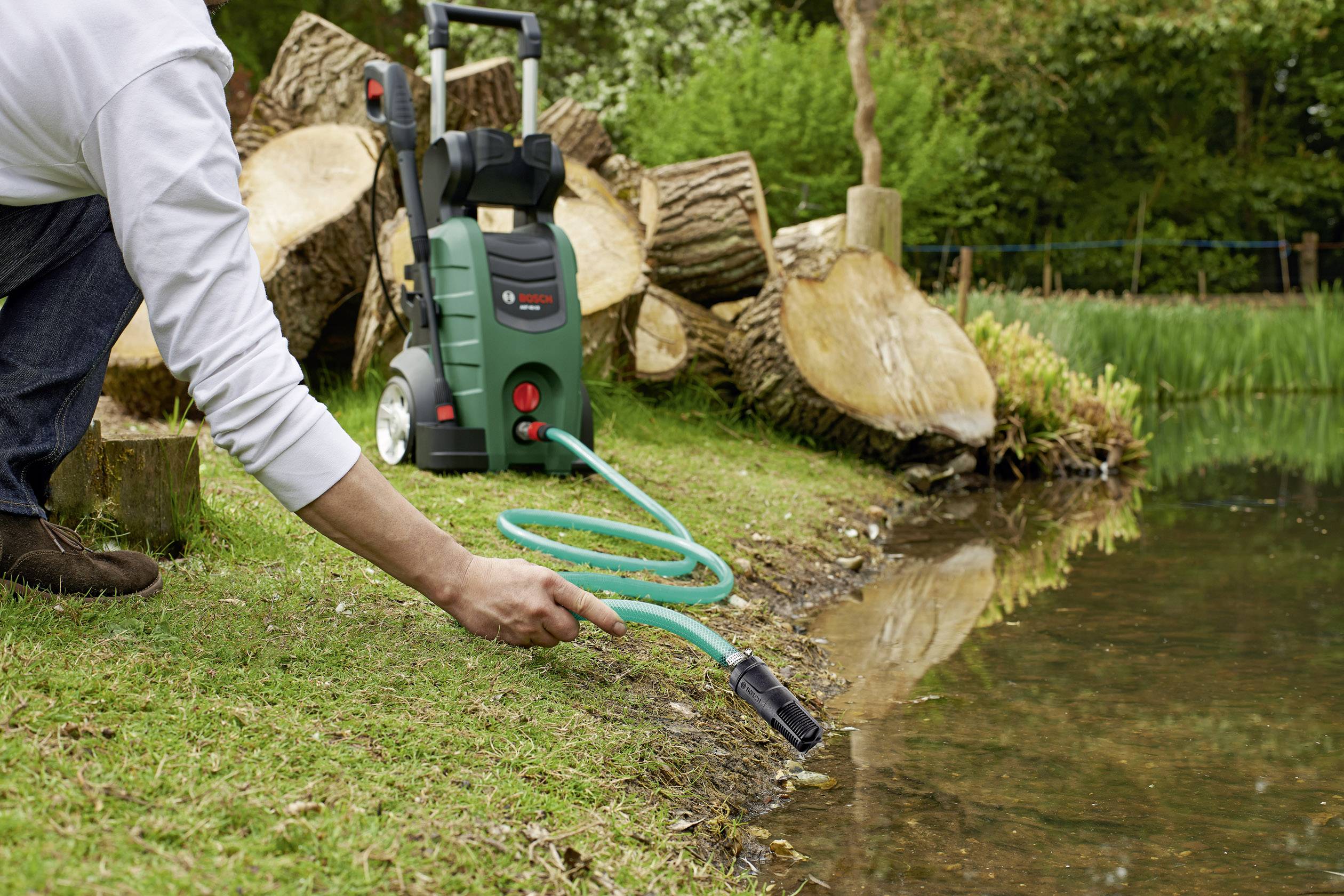 Person befüllt einen Hochdruckreiniger mit Wasser aus einem Teich, umgeben von Baumstämmen und Gras in einer natürlichen Umgebung.
