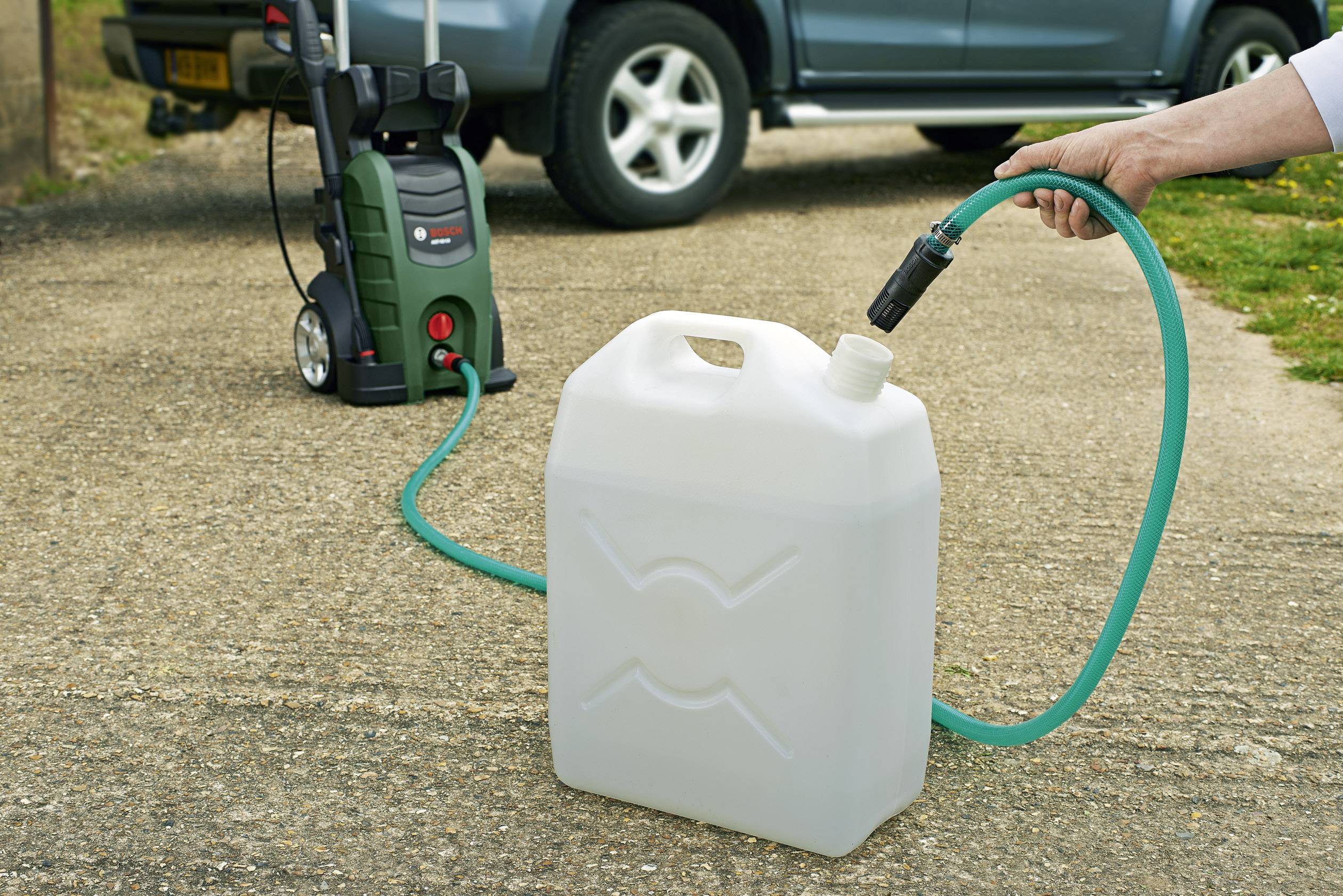 Eine Hand hält einen grünen Wasserschlauch, der an einen weißen Kanister angeschlossen ist. Im Hintergrund steht ein Hochdruckreiniger.