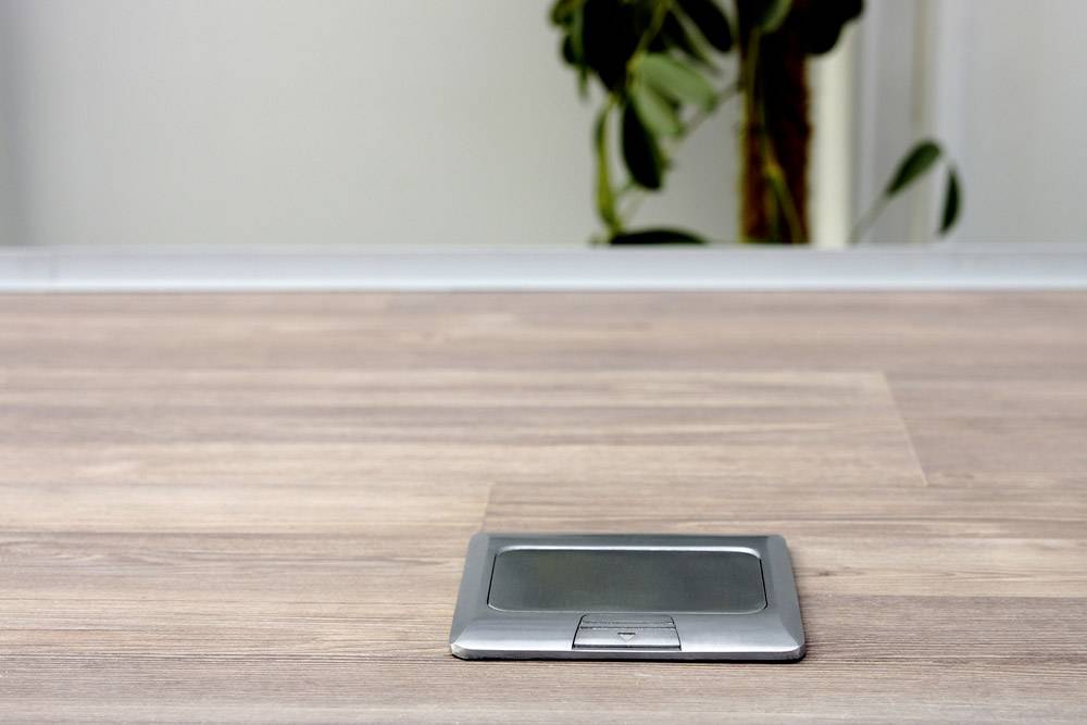 'Schreibtischoberfläche aus Holz mit eingebauter Steckdose.' Im Hintergrund ist eine Pflanze verschwommen sichtbar.