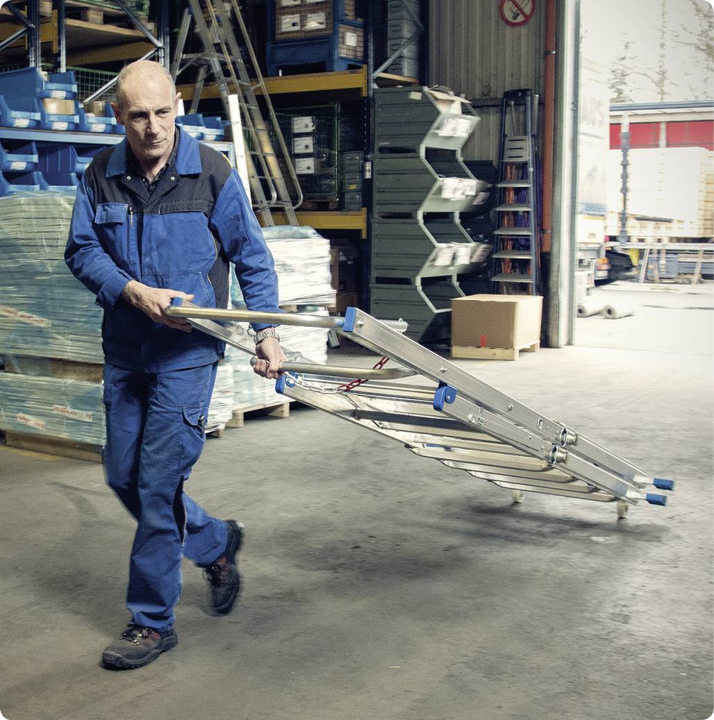 Ein Mann in Arbeitskleidung trägt eine zusammengeklappte Leiter durch ein Lagerhaus. Im Hintergrund sind Regale und Kartons zu sehen.