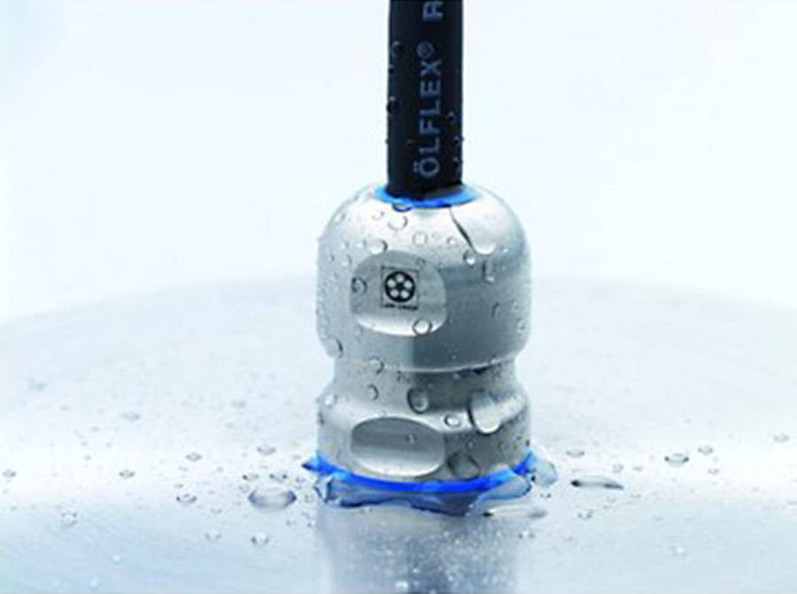 Ein rundes, silberfarbenes Kabelverschraubungselement, umgeben von Wassertropfen auf einer glatten, grauen Oberfläche.