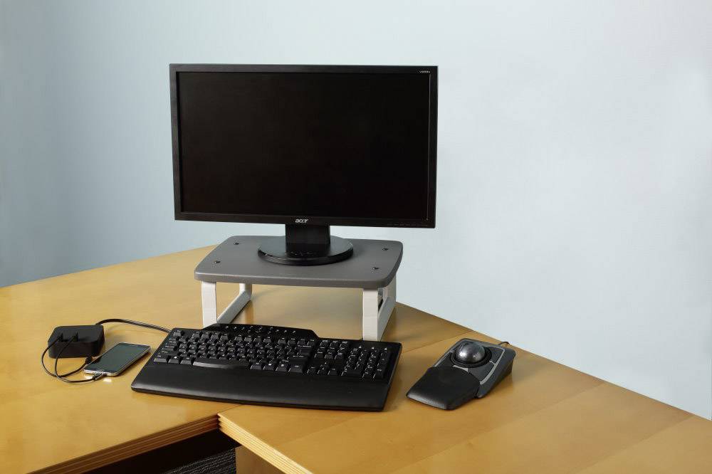Ein Computerarbeitsplatz mit Monitor auf einem Ständer, Tastatur, Trackball und Smartphone, verbunden durch ein Ladekabel.
