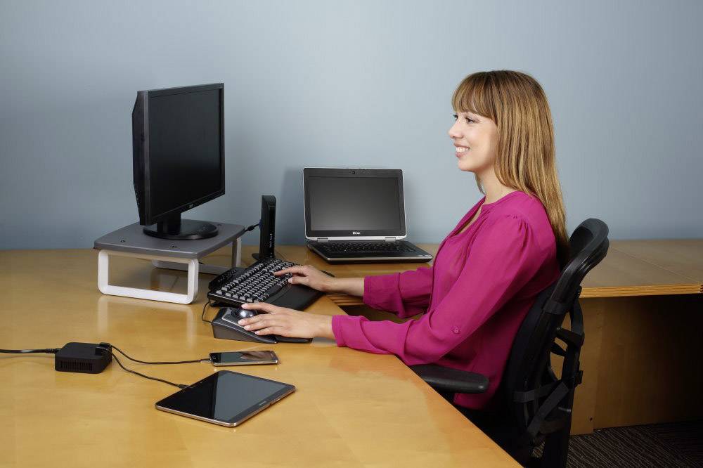 Eine Frau sitzt an einem Schreibtisch mit zwei Bildschirmen, einem Laptop, einem Tablet und einem Smartphone, lächelnd beim Arbeiten.