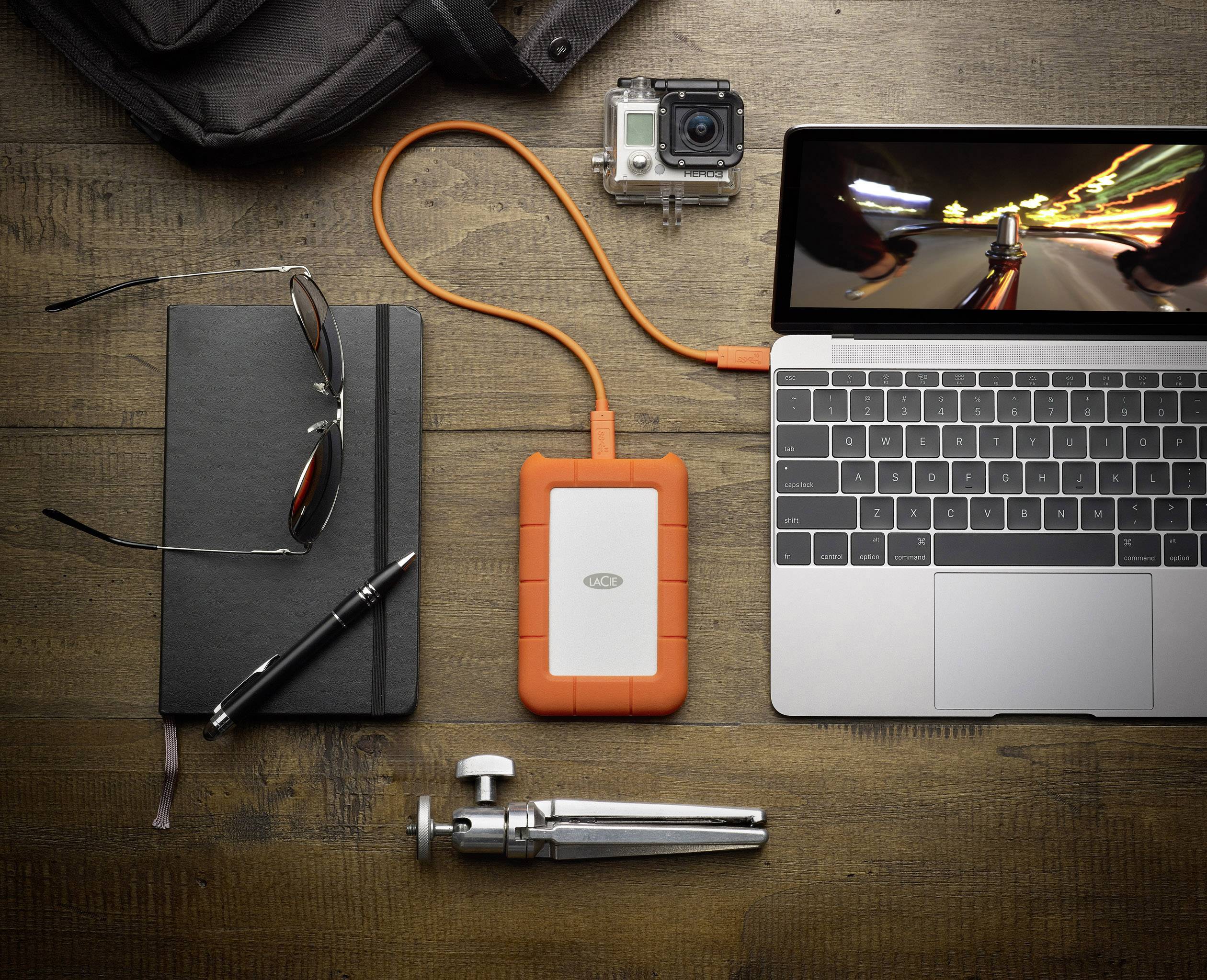 Externe Festplatte angeschlossen an Laptop auf Holztisch. Neben Laptop liegen Notizbuch, Brille, Stift, Kamera und kleines Stativ.