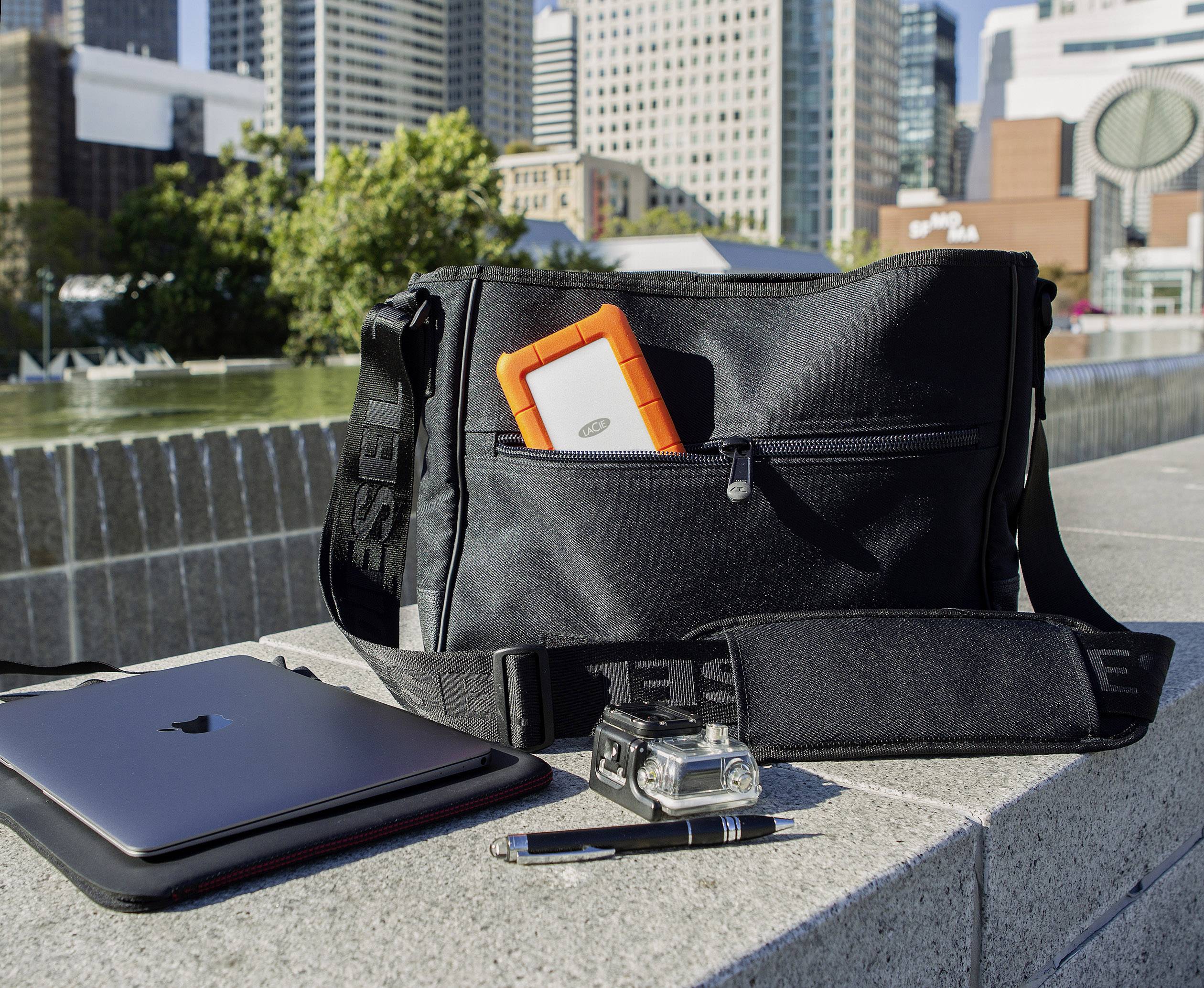 Eine schwarze Tasche mit oranger Festplatte, einem Laptop, einer Kamera und einem Stift in einer städtischen Umgebung im Freien.