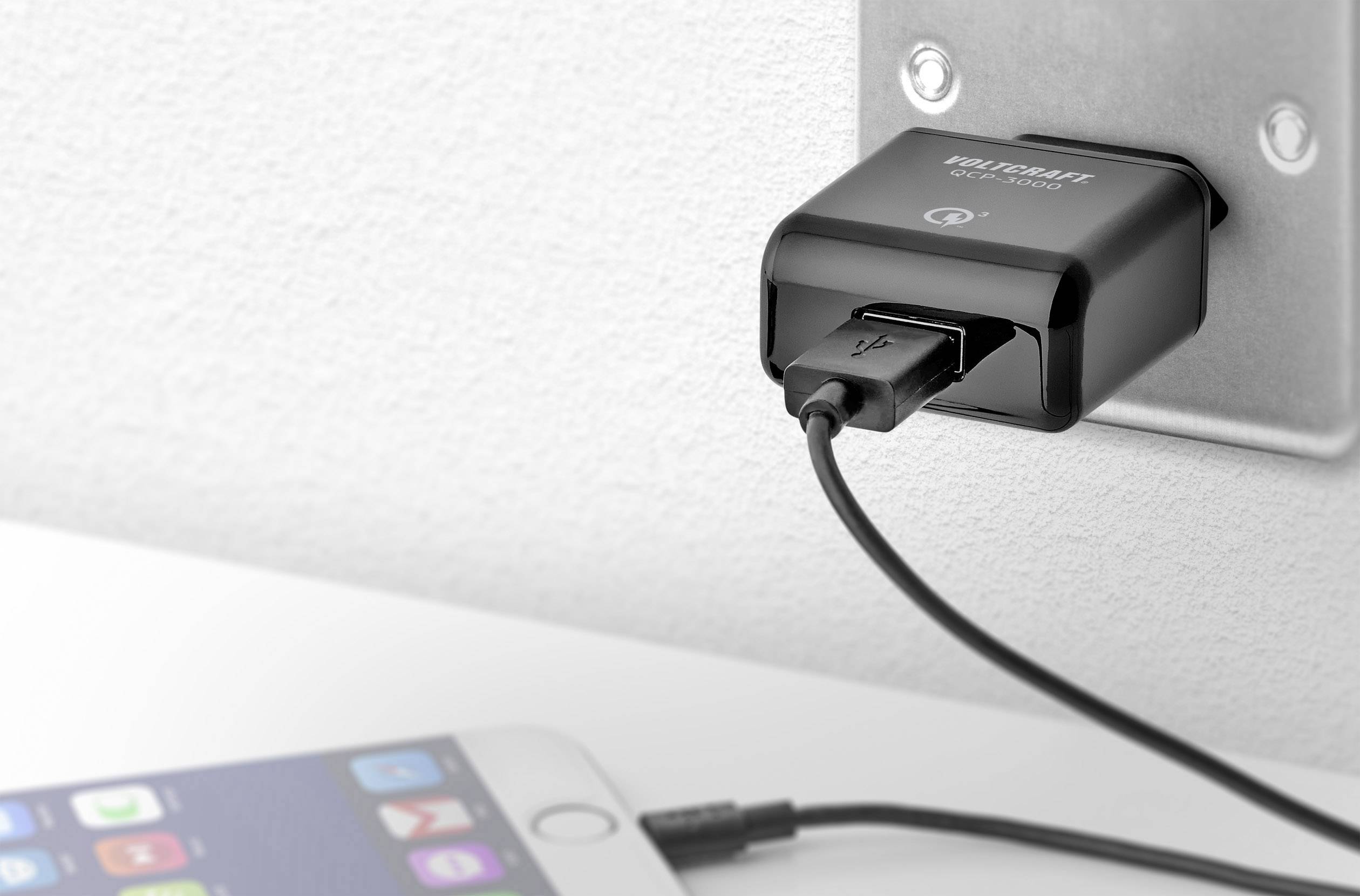 Ein schwarzes USB-Ladegerät ist an eine Wandsteckdose angeschlossen und lädt ein Smartphone, das auf einer weißen Oberfläche liegt.