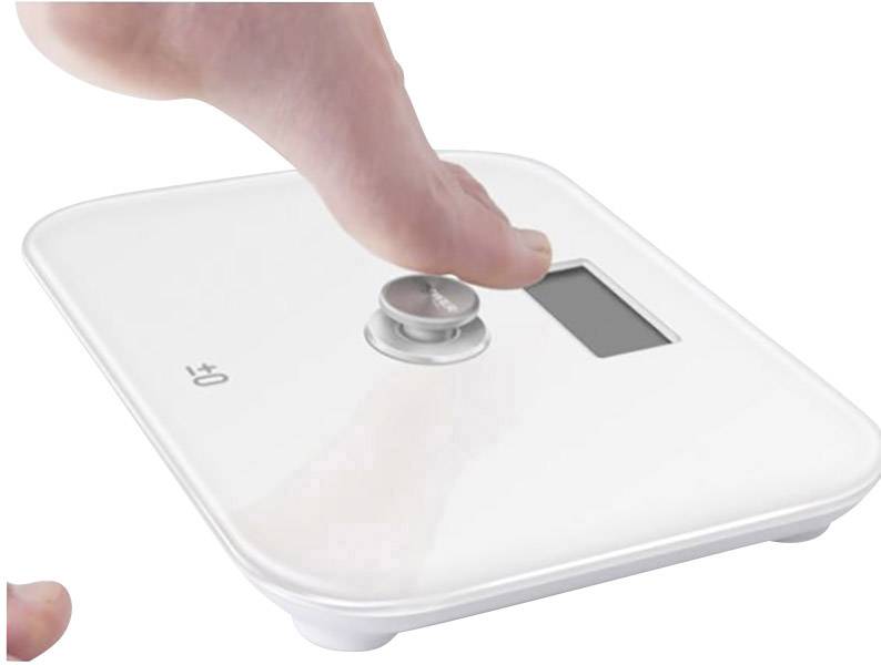 Ein nackter Fuß betätigt eine digitale Waage mit einem weißen Gehäuse und einem Display, das Gewicht in Kilogramm anzeigt.