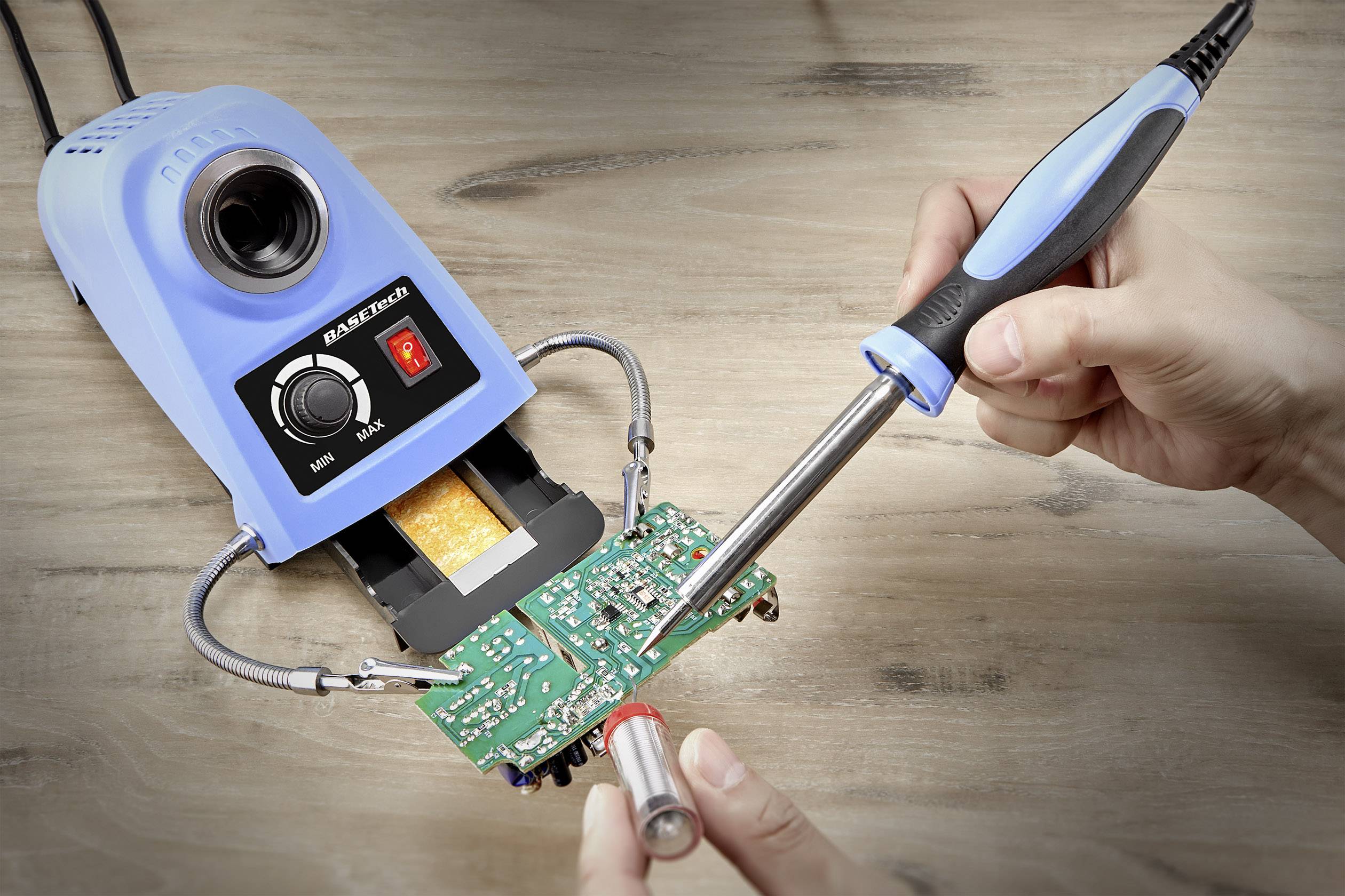 Ein Lötkolben wird benutzt, um eine elektronische Schaltung auf einer grünen Platine zu löten; eine helle Lötstation ist im Hintergrund.