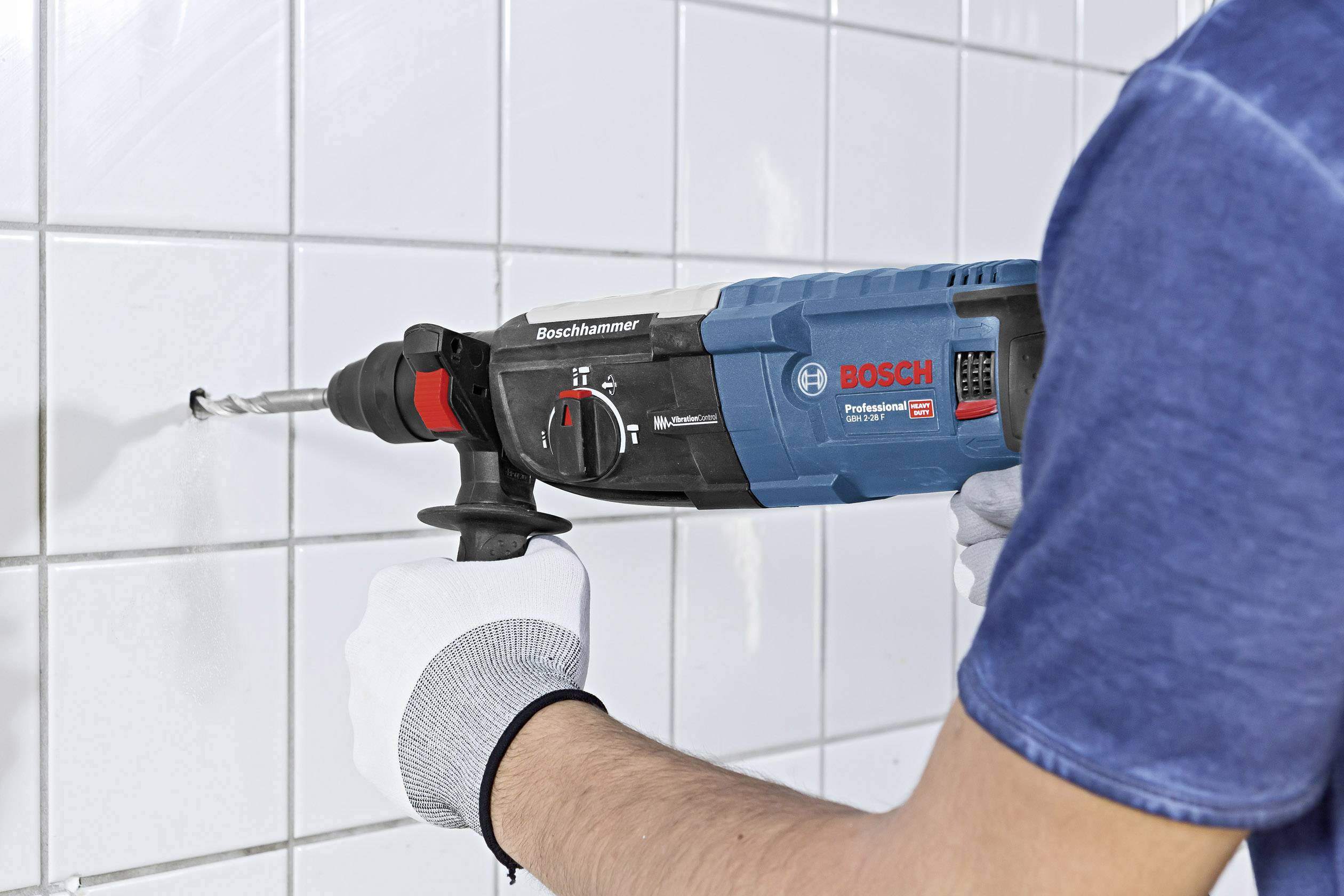 Person bohrt mit einer blauen Bosch-Bohrmaschine ein Loch in eine weiße Fliesenwand, trägt dabei weiße Handschuhe.