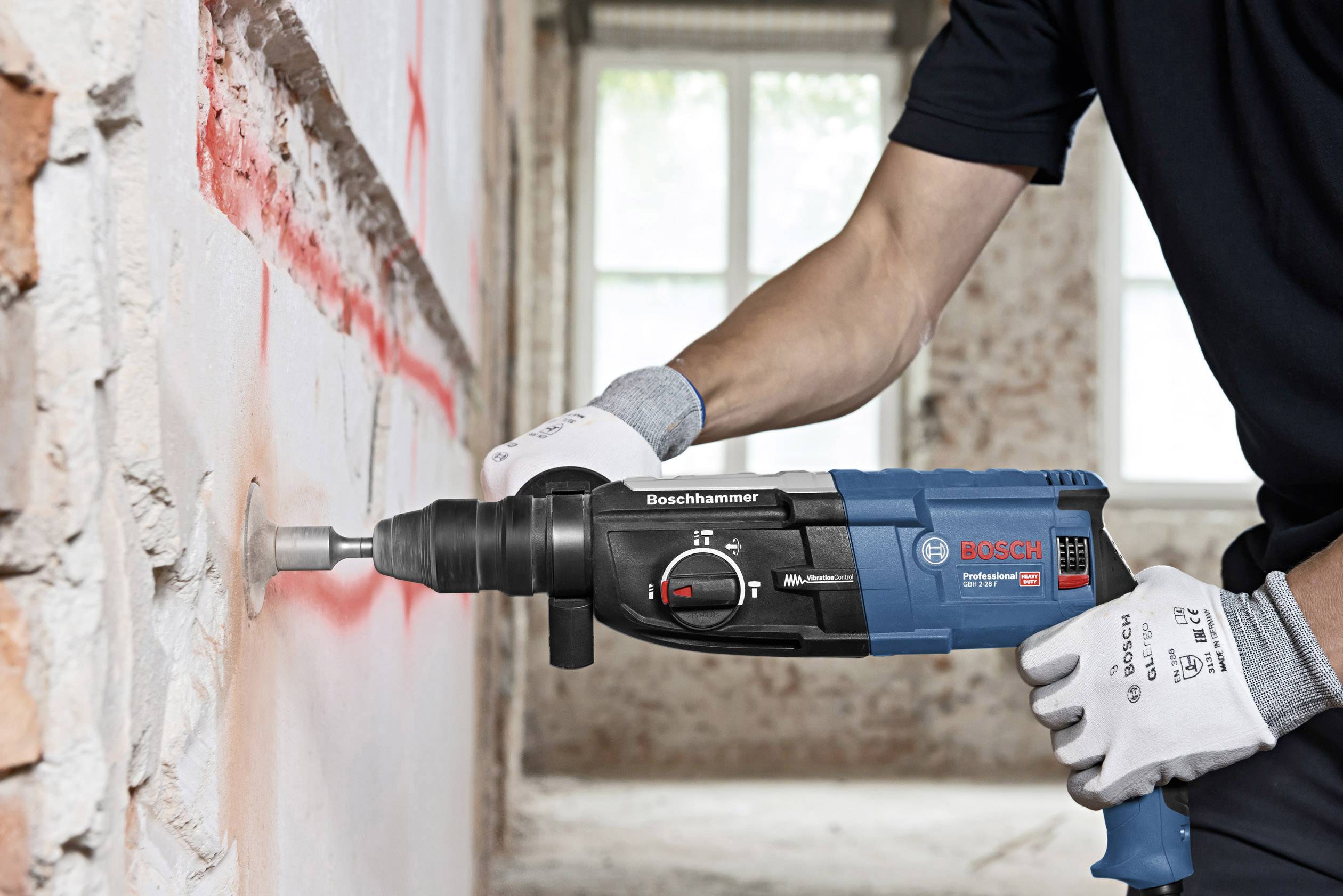 Eine Person verwendet einen BOSCH Bohrhammer, um in eine mit rotem Staub bedeckte Betonwand zu bohren. Handschuhe schützen die Hände.