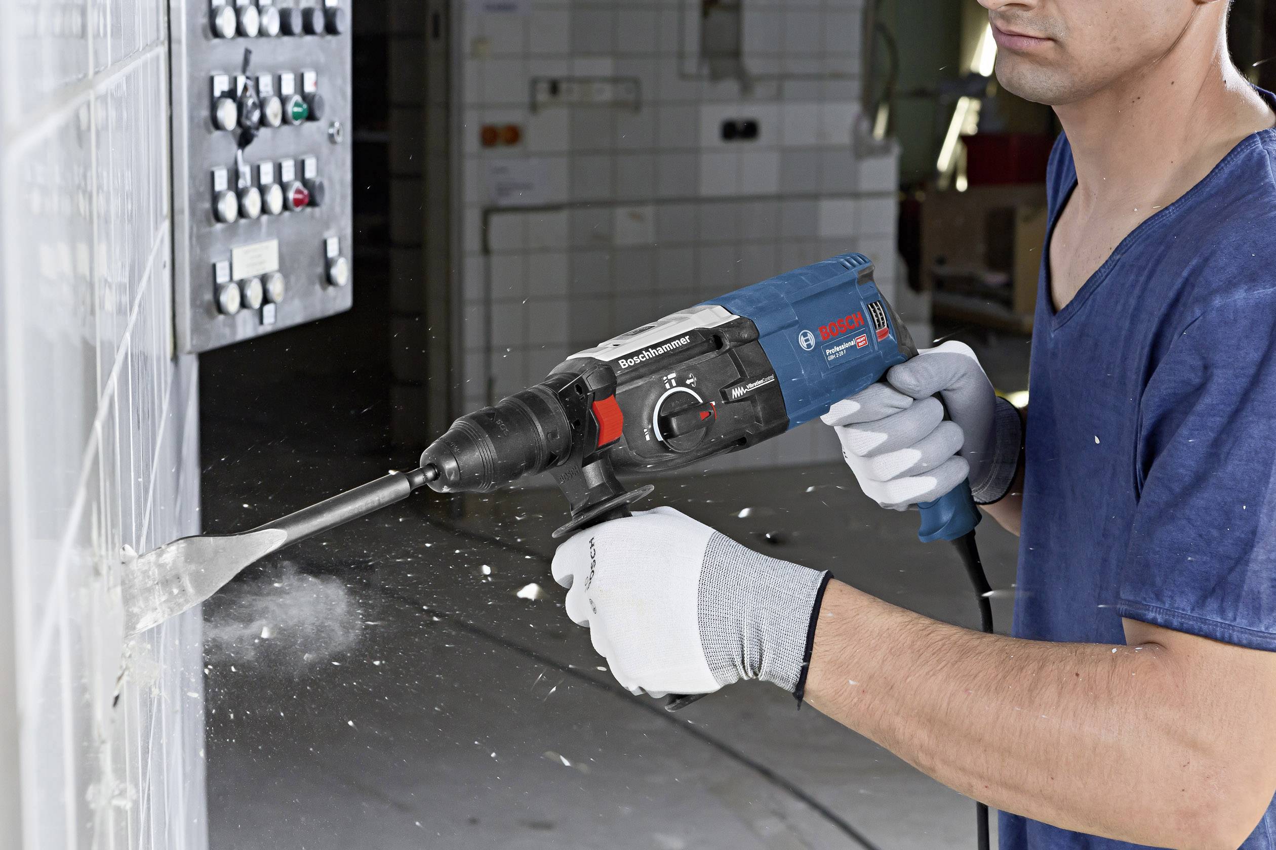 Eine Person im blauen Hemd und grauen Handschuhen arbeitet mit einem Bohrhammer an einer Wand in einem industriellen Raum.