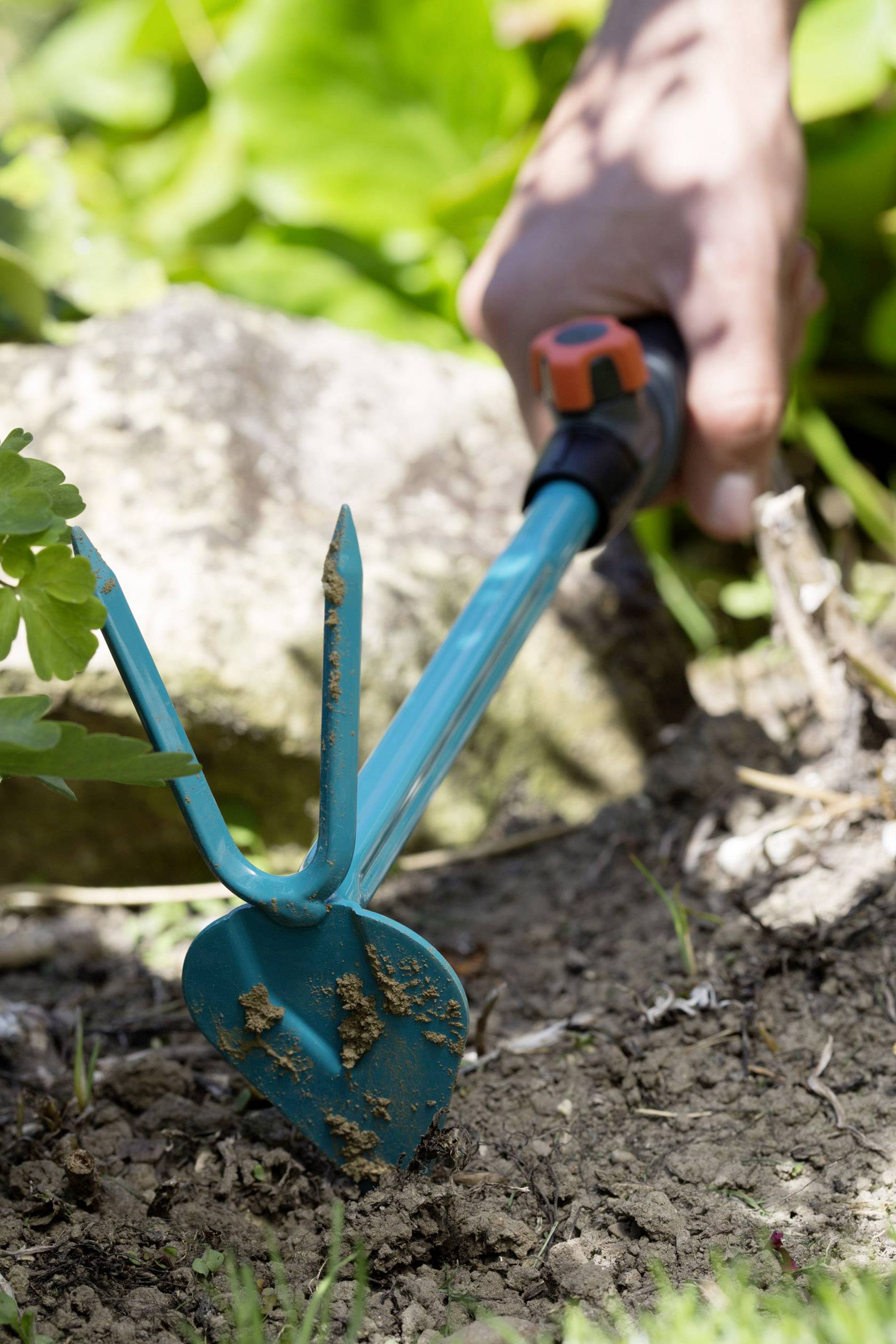 Eine Hand hält ein Gartengerät, das Erde in einem Beet bearbeitet. Im Hintergrund sieht man unscharfe Blätter.