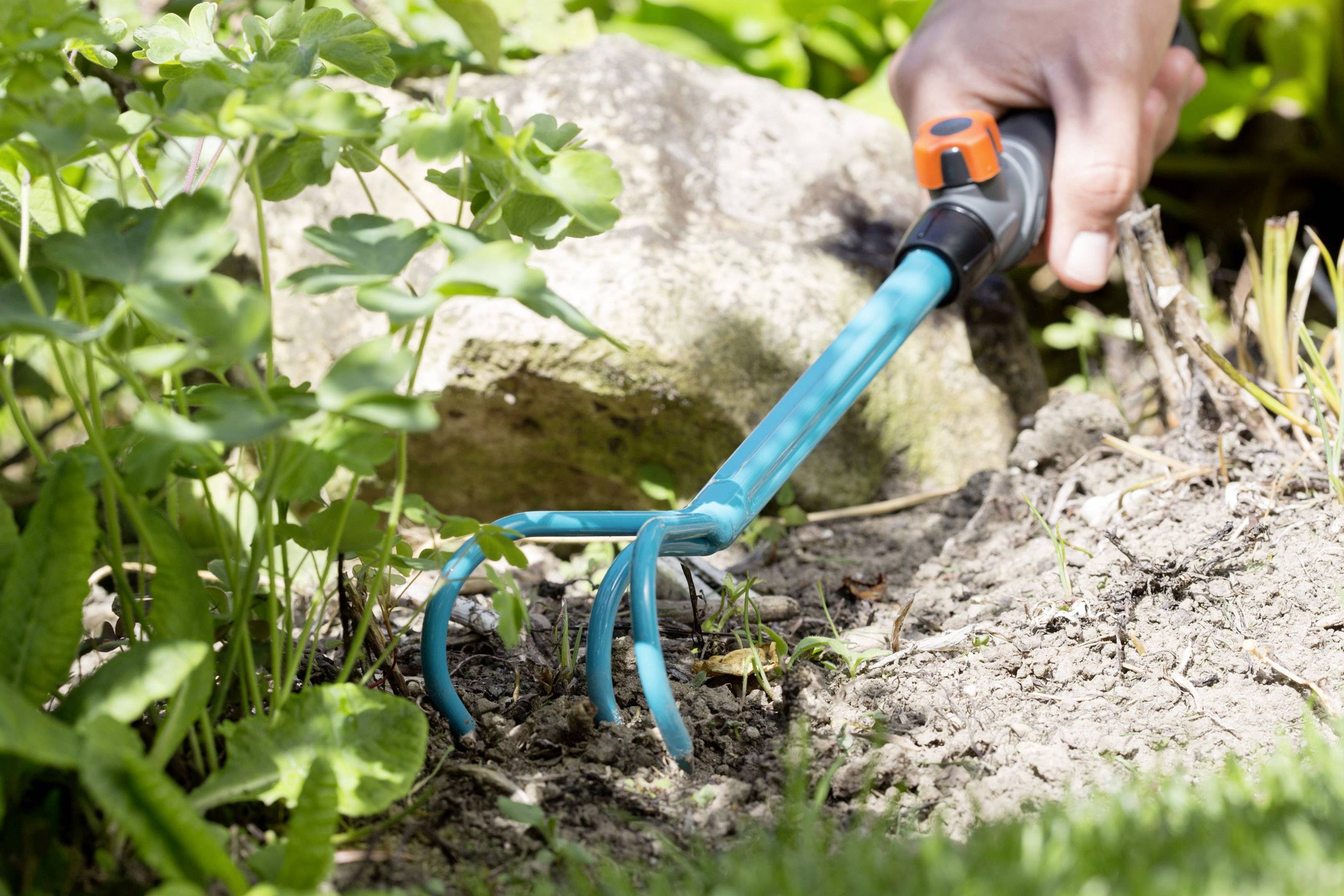 Eine Hand hält eine Gartengeräte-Hacke und bearbeitet damit Erde in einem Garten. Pflanzen und Steine sind im Hintergrund sichtbar.