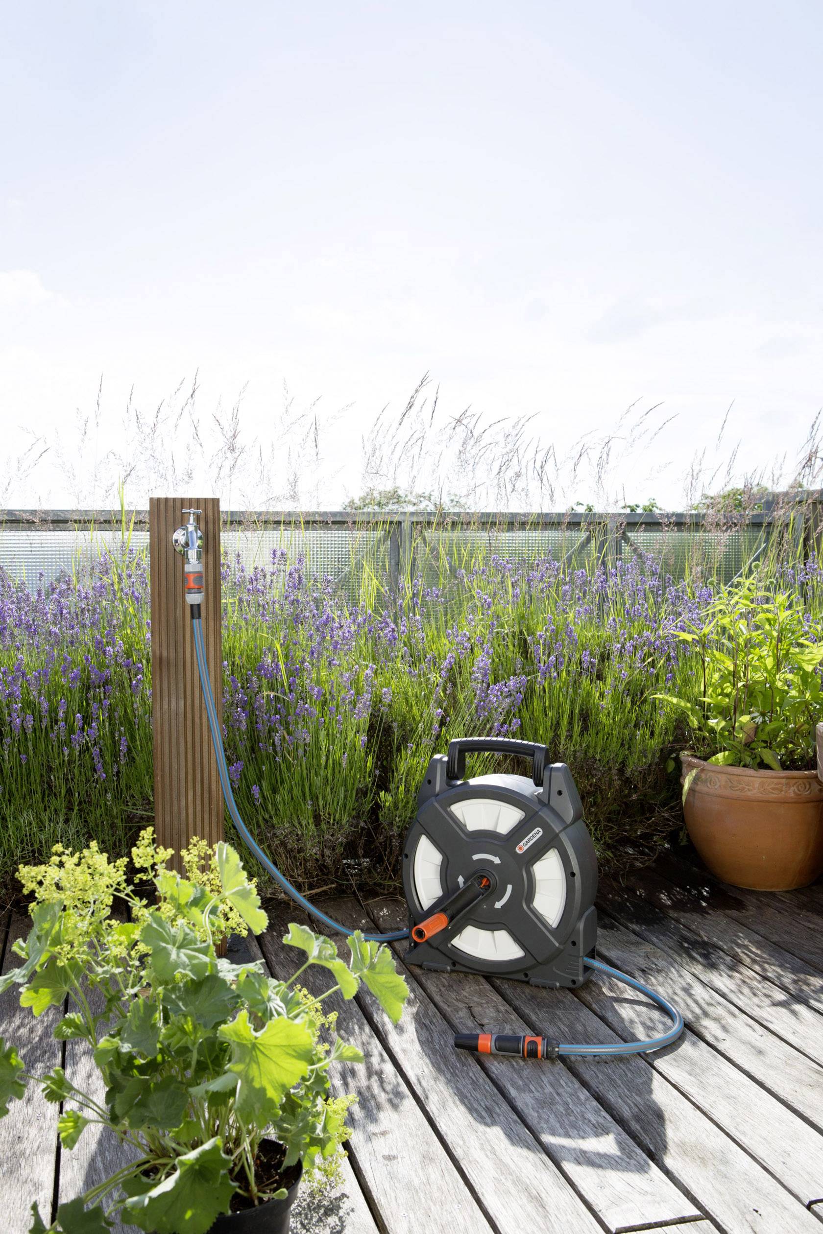 Ein Wasserschlauch auf einer Holzterrasse, umgeben von blühenden Pflanzen und Lavendel im Hintergrund, an einem sonnigen Tag.