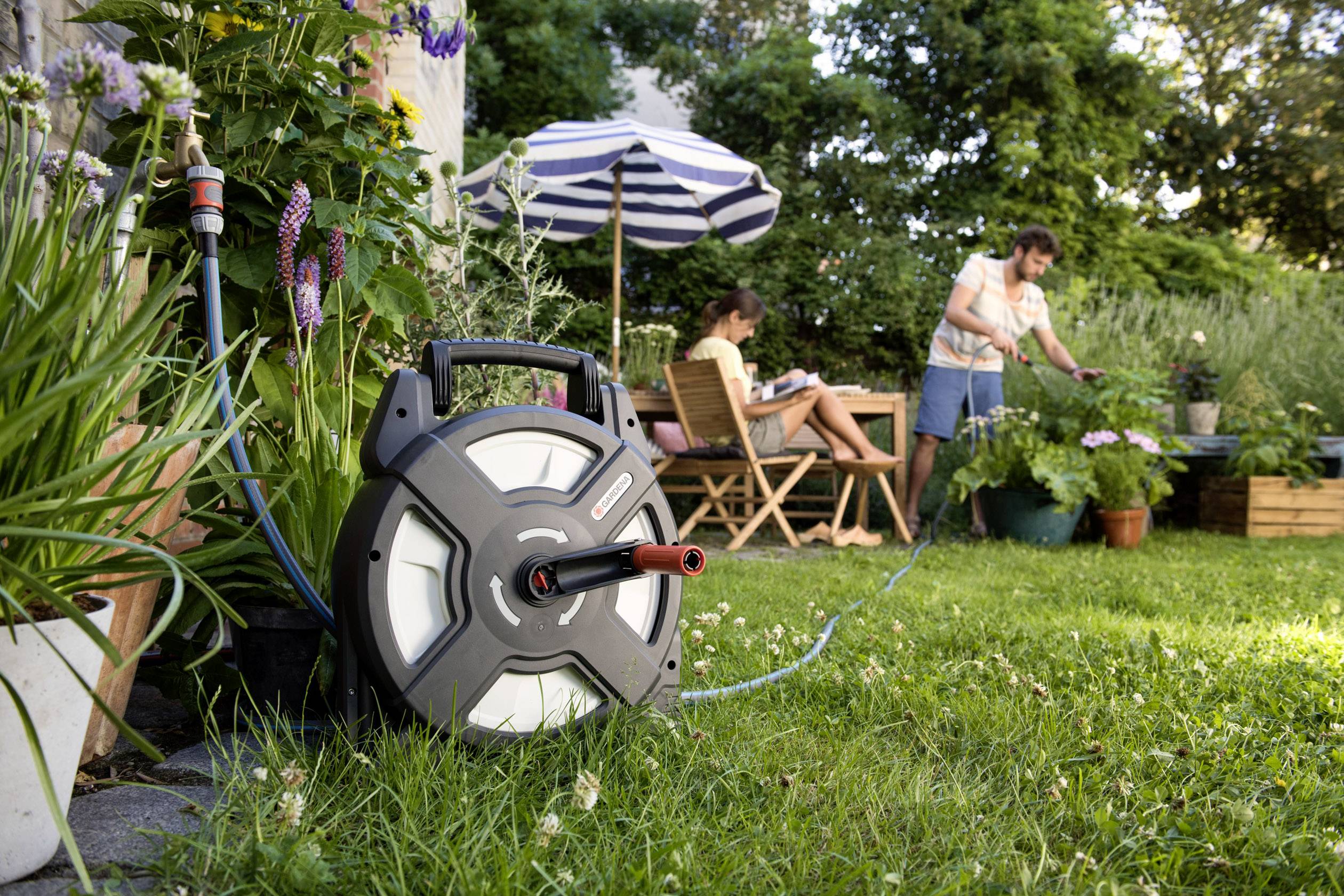 Gartenszene mit einem Mann, der Pflanzen gießt, während eine Frau auf einem Stuhl liest; im Vordergrund ein Wasserschlauch auf einer Rolle.