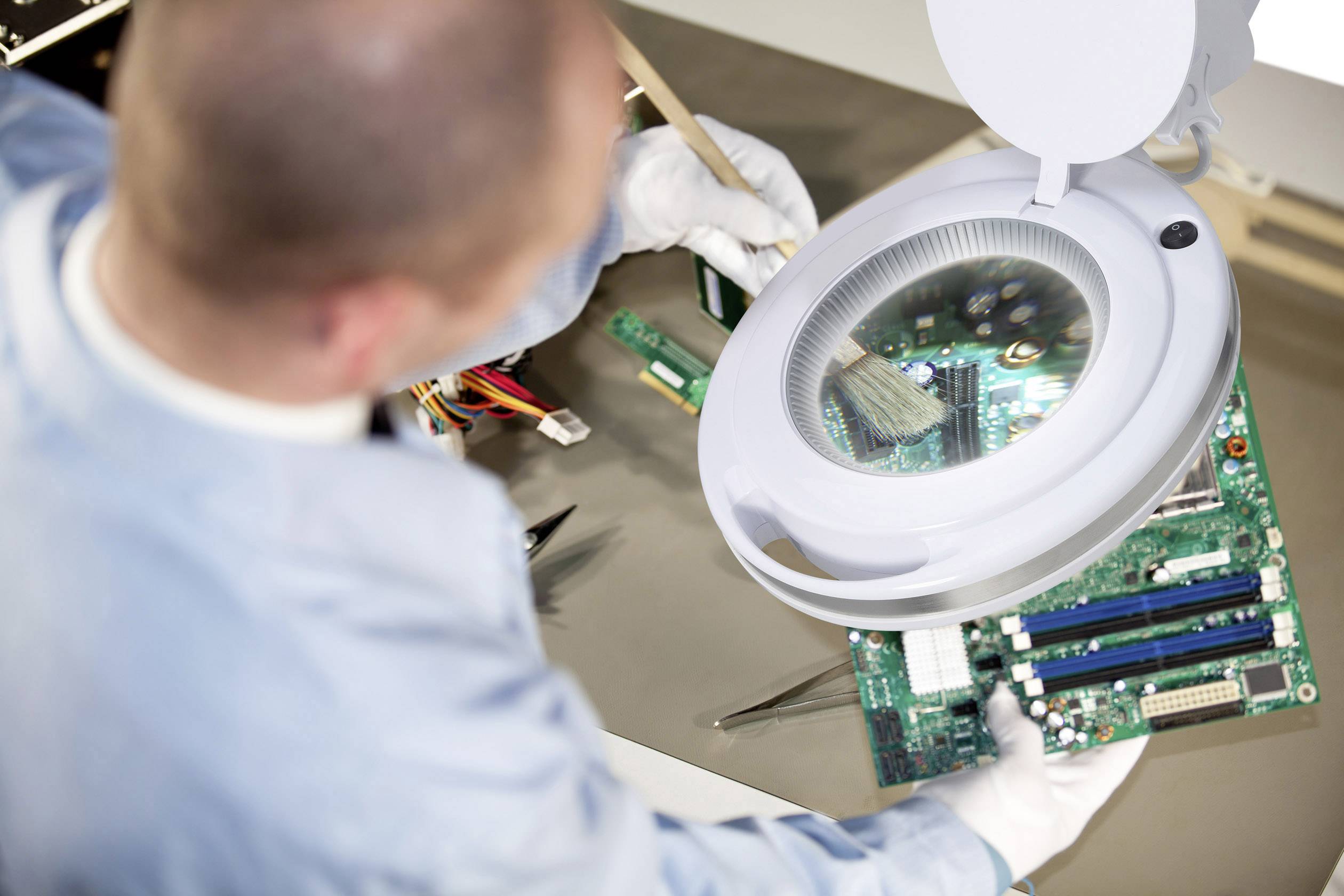 Ein Techniker reinigt eine Computerplatine unter einer beleuchteten Lupe mit einem Pinsel.