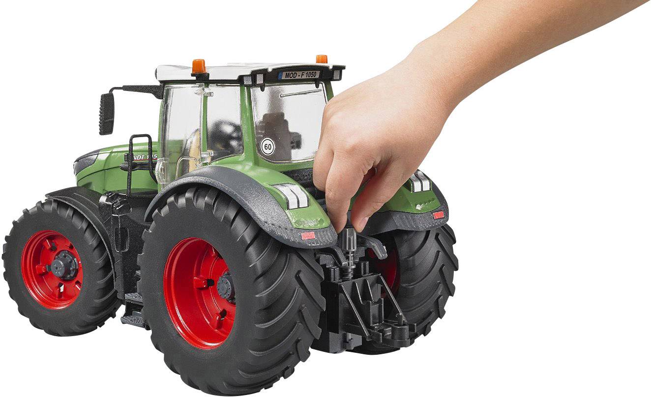 Eine Hand justiert ein Modell eines grünen Traktors mit großen schwarzen Reifen und rotem Inneren auf einer weißen Hintergrundfläche.