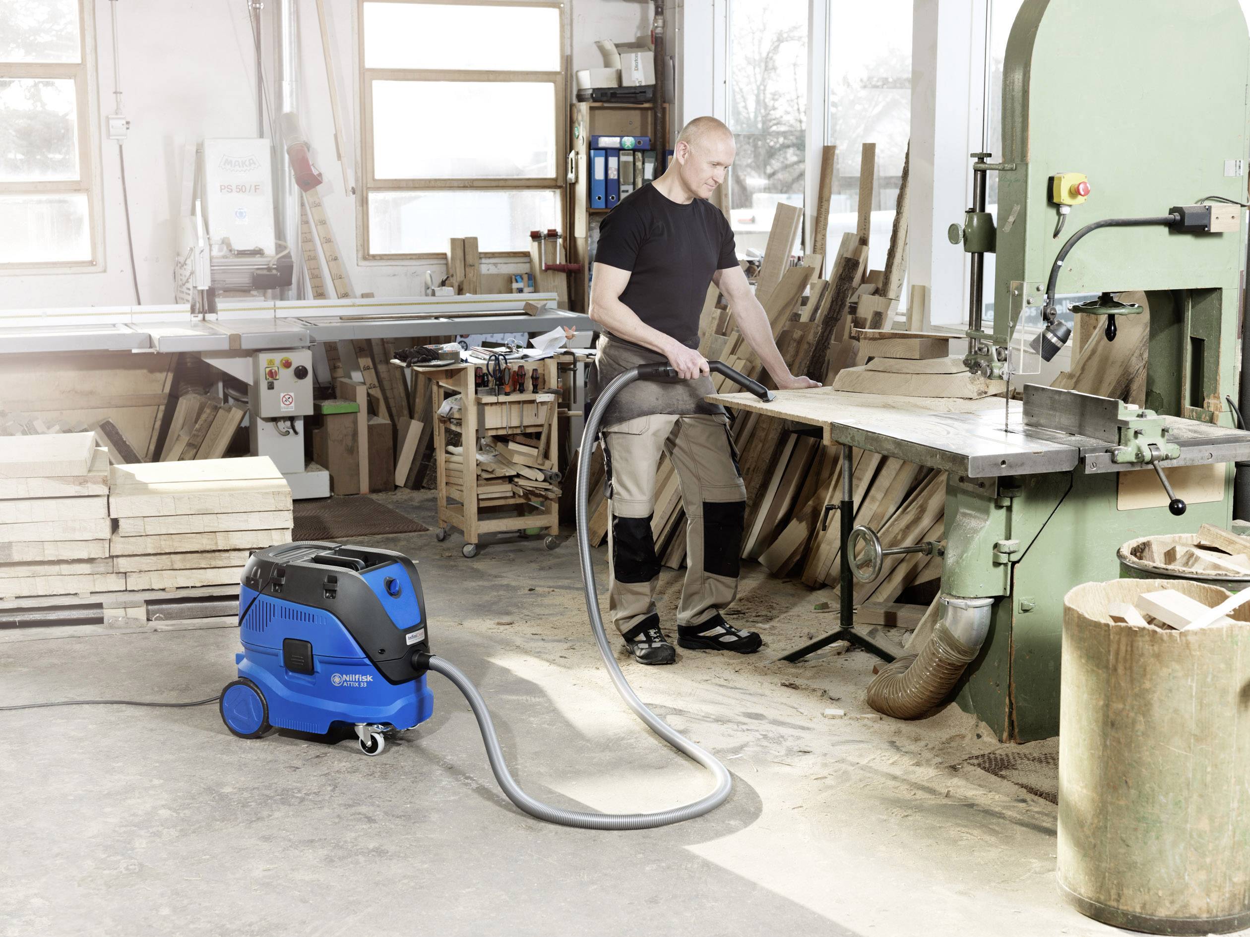 Ein Mann in einer Werkstatt arbeitet mit einer Bandsäge. Der Boden ist sauber, da ein blauer Industriesauger den Holzstaub absaugt.