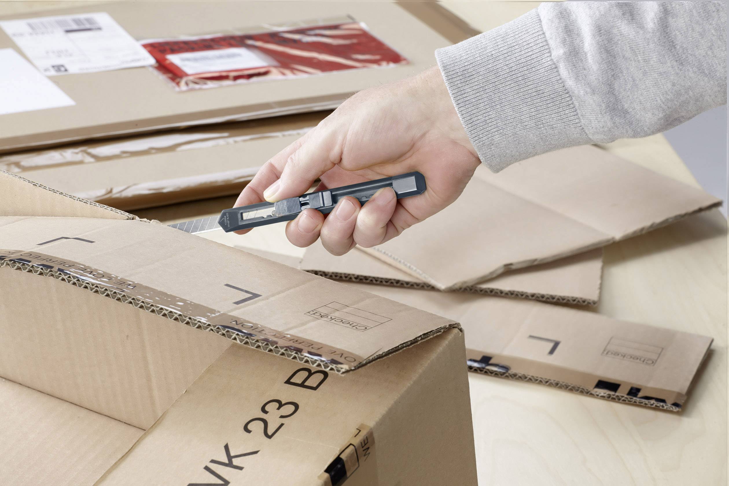 Eine Hand verwendet ein Teppichmesser, um einen Pappkarton zu öffnen. Der Karton liegt auf einem Tisch und ist teilweise geöffnet.