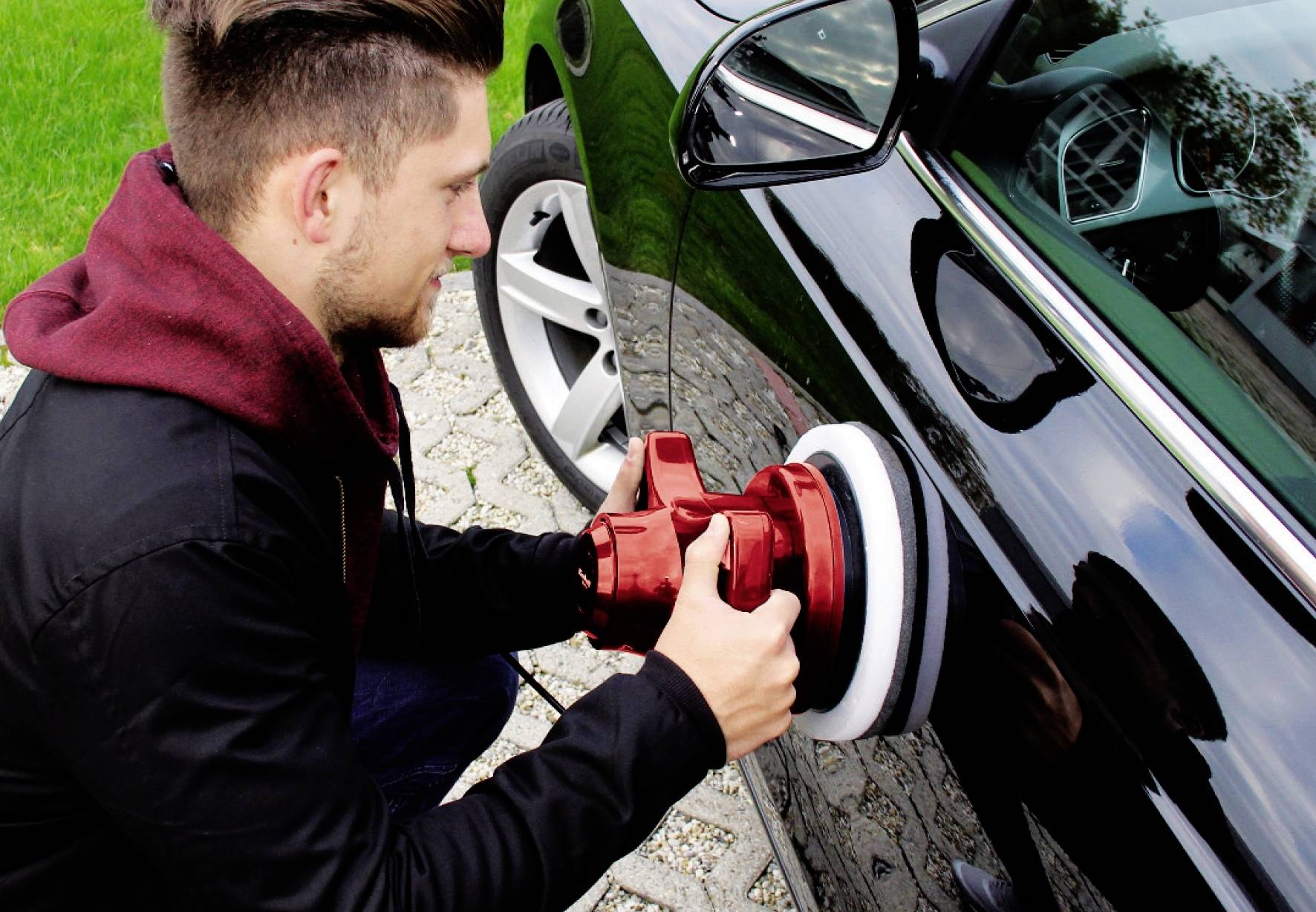 Mann poliert ein Auto mit einer roten Poliermaschine, während er sich auf dem Bürgersteig neben einem schwarzen Auto befindet.