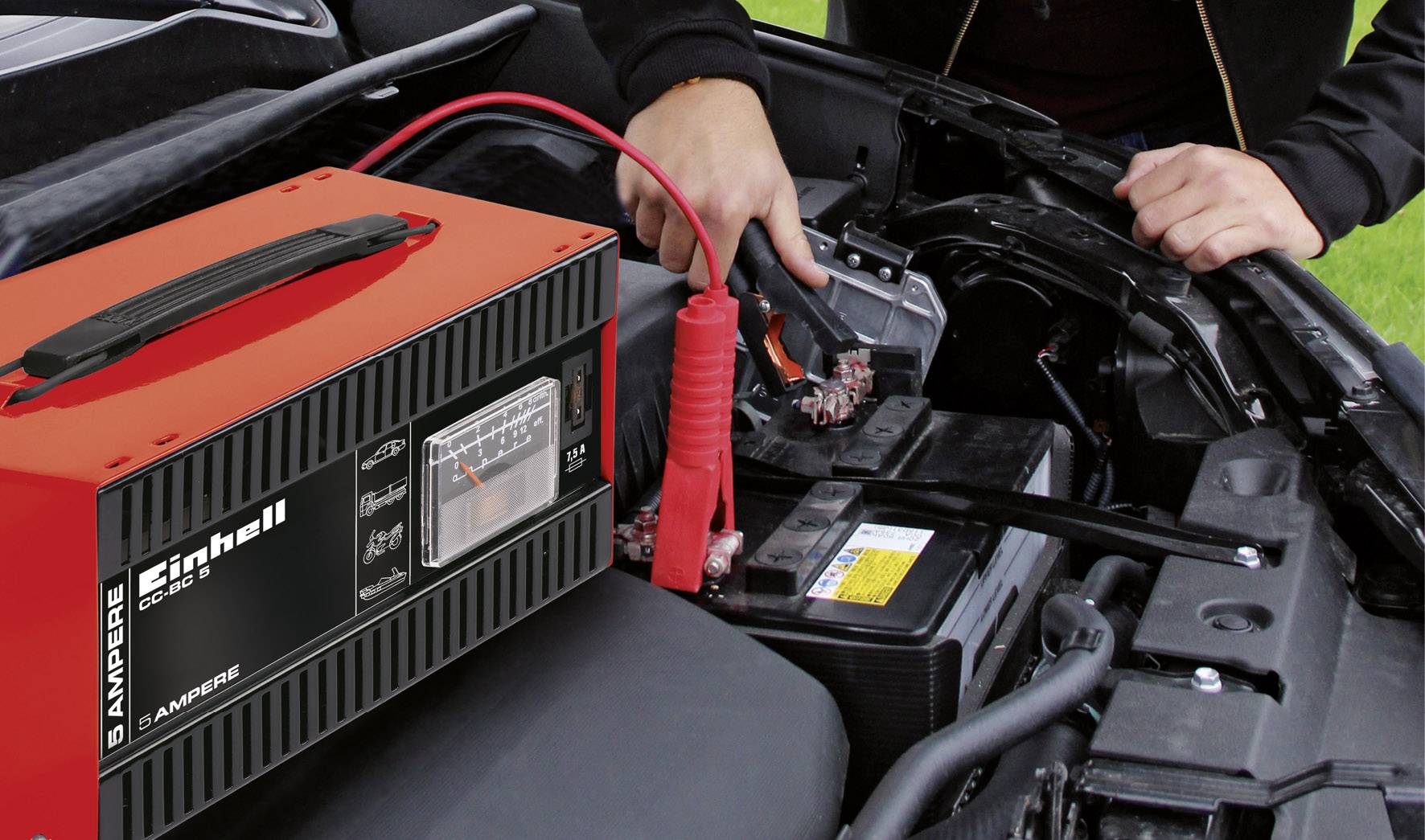 Eine Person lädt eine Autobatterie mit einem Ladegerät auf. Das Ladegerät zeigt 6 Ampere an. Kabel sind an der Batterie angeschlossen.