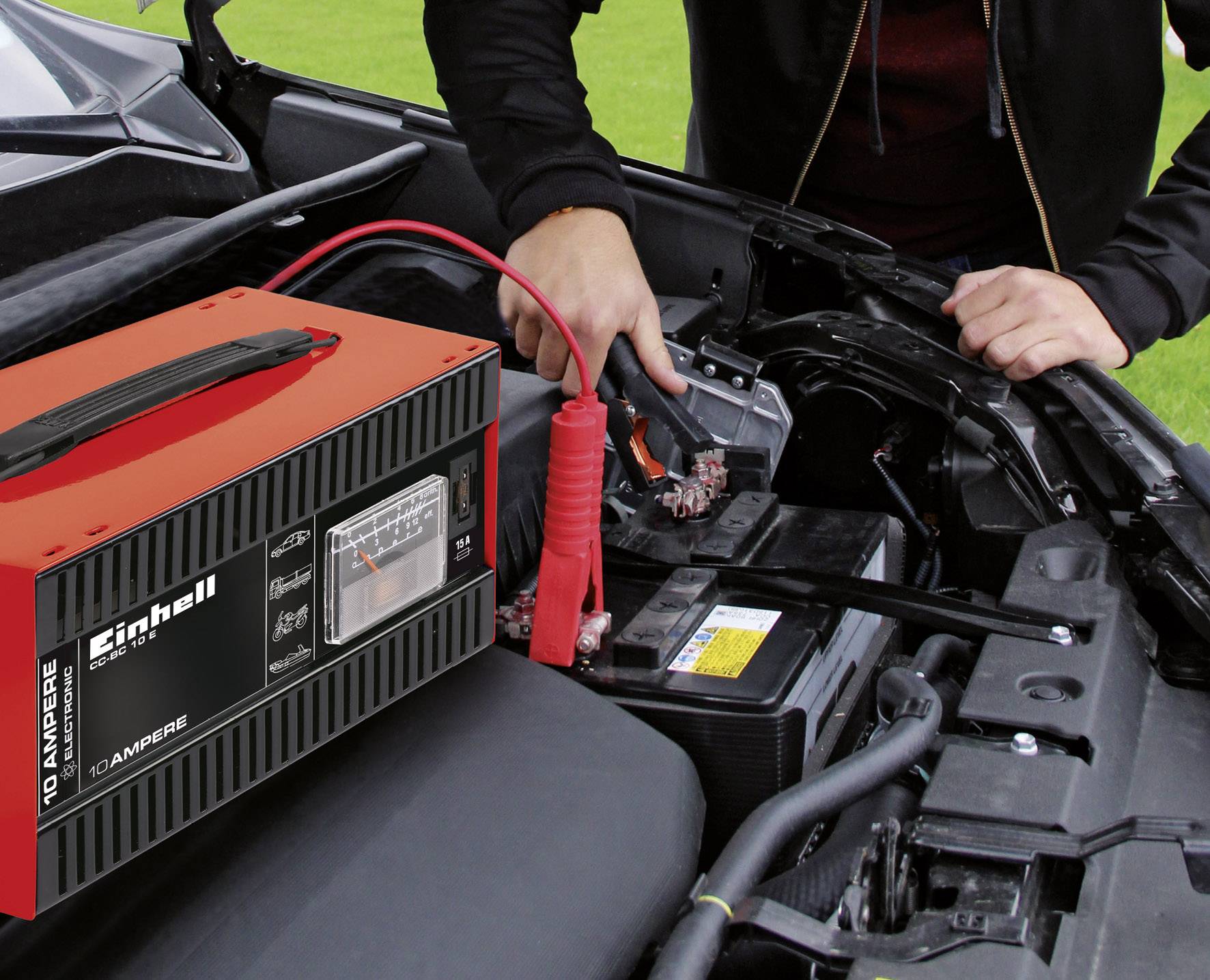 Person überbrückt Autobatterie mit Starterkabeln, rotes Ladegerät im Vordergrund. Im Hintergrund eine motorhaubenoffene Wiese.