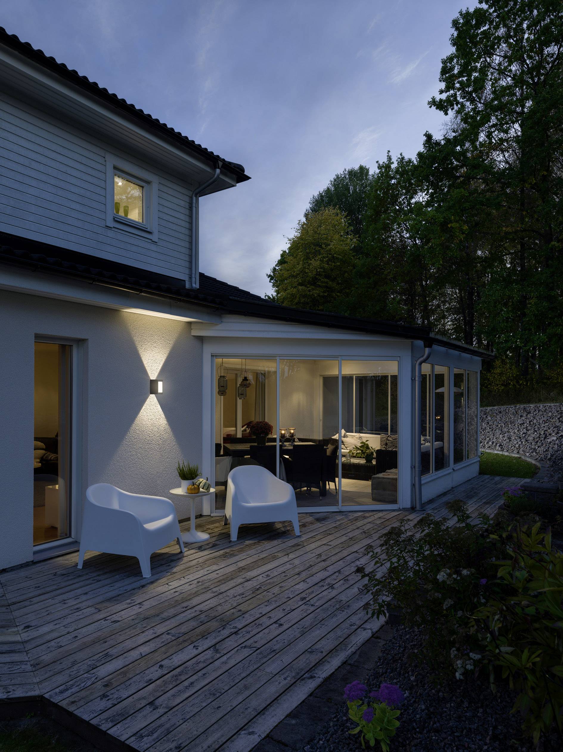 Moderner Wintergartenanbau an ein Einfamilienhaus, umgeben von Bäumen, mit abendlicher Beleuchtung. Zwei weiße Stühle auf der Holzterrasse.