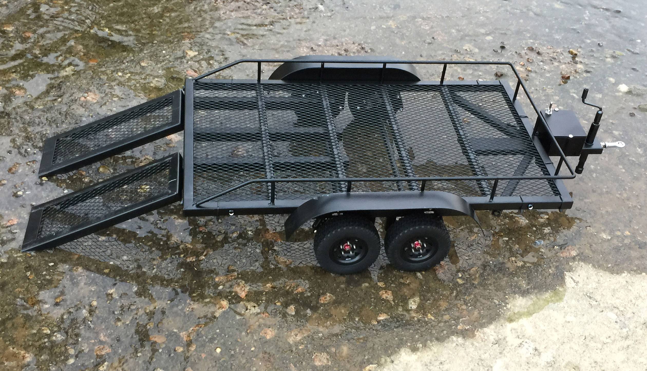 Ein schwarzer Anhänger mit zwei Achsen und abklappbaren Rampen steht auf einem steinigen Untergrund, umgeben von Wasser.