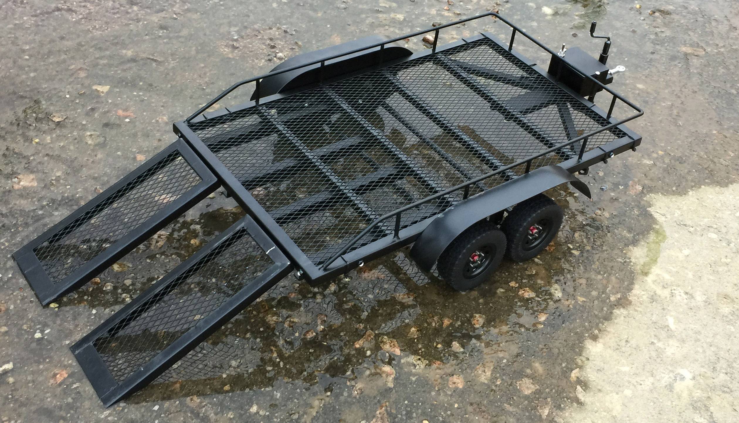 Ein schwarzer, zweiachsiger Anhänger mit geöffneten Auffahrrampen auf nassem Untergrund. Geeignet für den Transport von Fahrzeugen.