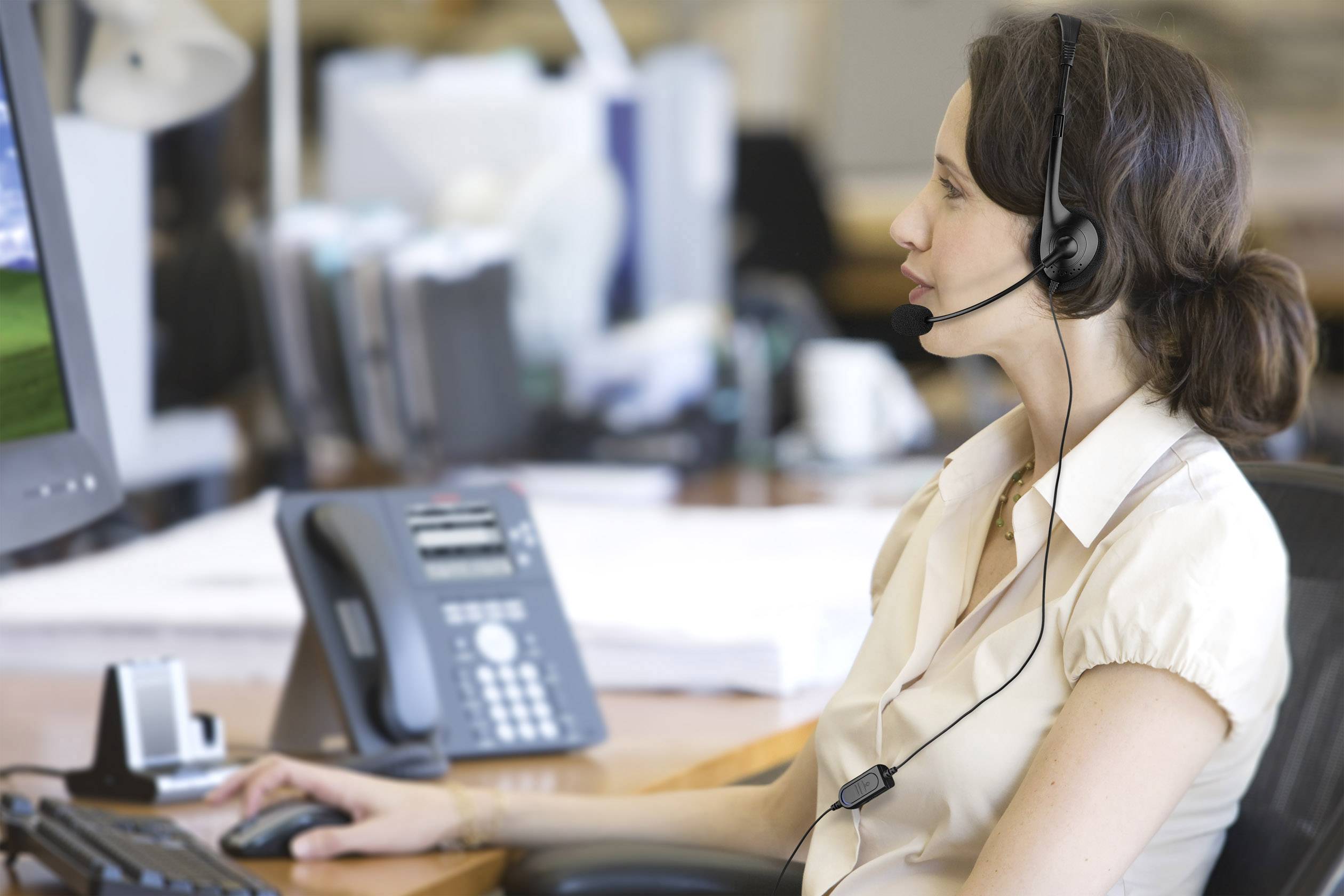 Eine Frau mit Headset arbeitet konzentriert am Computer in einem Büro. Im Hintergrund sind ein Tischtelefon und unscharfe Büroelemente zu sehen.