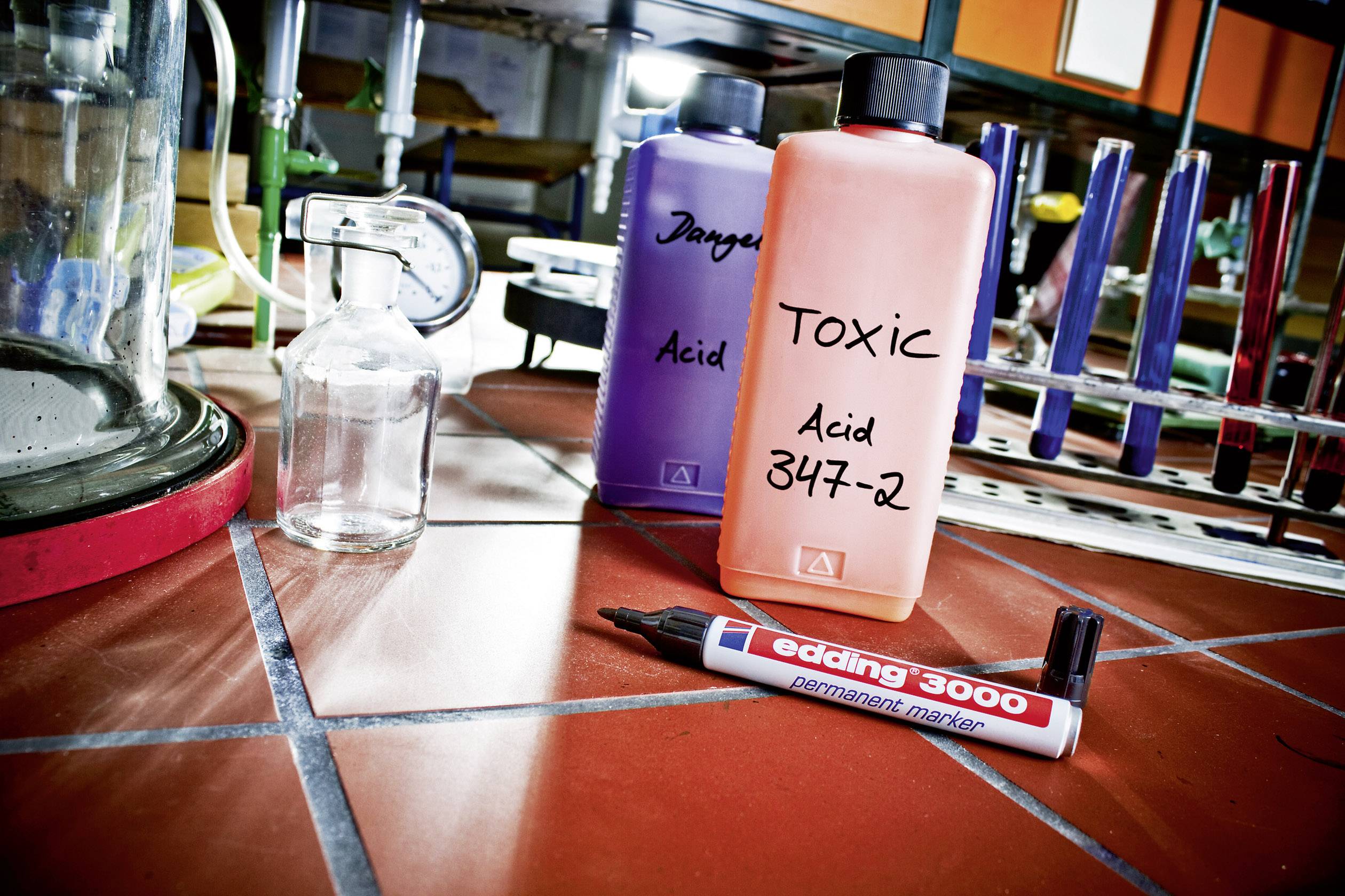 Zwei Flaschen mit Aufschrift 'Danger Acid' und 'Toxic Acid 347-2' stehen auf einem Fliesenboden in einem Labor neben einem Marker.