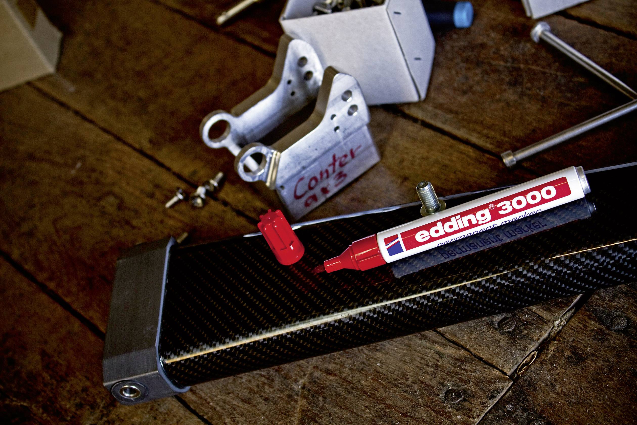 Ein roter Edding-Marker liegt auf einer Kohlefaser-Oberfläche, umgeben von Metallteilen auf einem Holztisch.